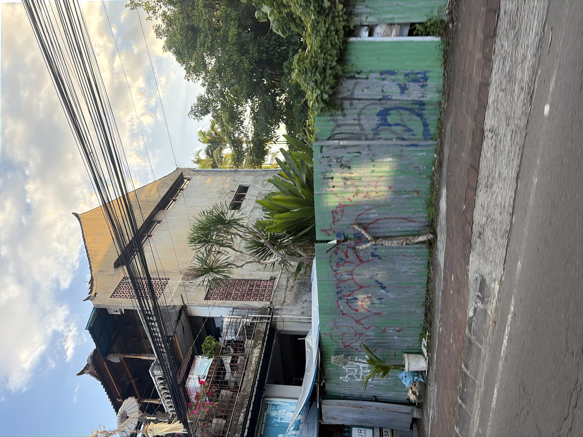 The image captures a street view in the Ubud District, Indonesia, featuring a multi-story building with traditional Balinese architectural elements on the left, characterized by its layered rooflines and ornate decorations. A balcony adorned with plants and an umbrella is visible on the second level. The building's facade is a light color, with some sections appearing weathered. A series of black electrical wires stretch across the upper portion of the frame, obscuring parts of the sky and the building.
In the foreground, a corrugated metal fence, painted in shades of green and exhibiting graffiti, runs along the side of the street. This fence is partially overgrown with vines and other greenery. A small potted plant stands near the fence. A paved sidewalk is present between the fence and the road, which appears to be asphalt. Lush green trees and foliage are visible behind the fence and to the right of the building, suggesting a tropical environment. The lighting indicates it might be late afternoon, with the sun casting a warm glow on the building. No people are clearly visible in the scene.