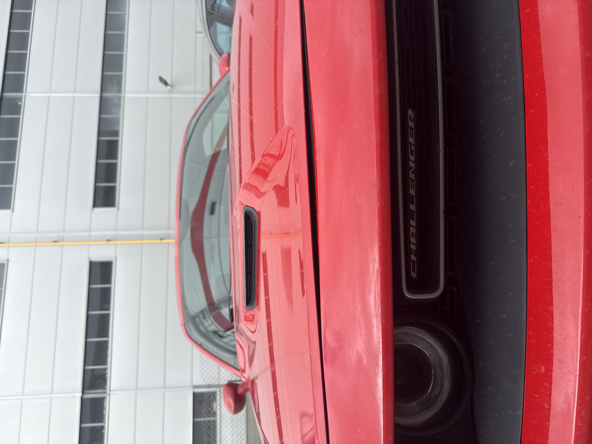A partial view of a red Dodge Challenger, positioned vertically, fills the majority of the frame. Visible features include the driver's side door with an air intake, part of the front windshield, the black panel behind the front tire with the word "CHALLENGER" embossed on it, a portion of the front tire, and the vehicle's red side panels. The car is parked next to a gray building with multiple rectangular windows. A yellow horizontal pipe runs across the building's facade. The location is unspecified, and there is no mention of Sofia, Bulgaria in the image. There are no people visible.