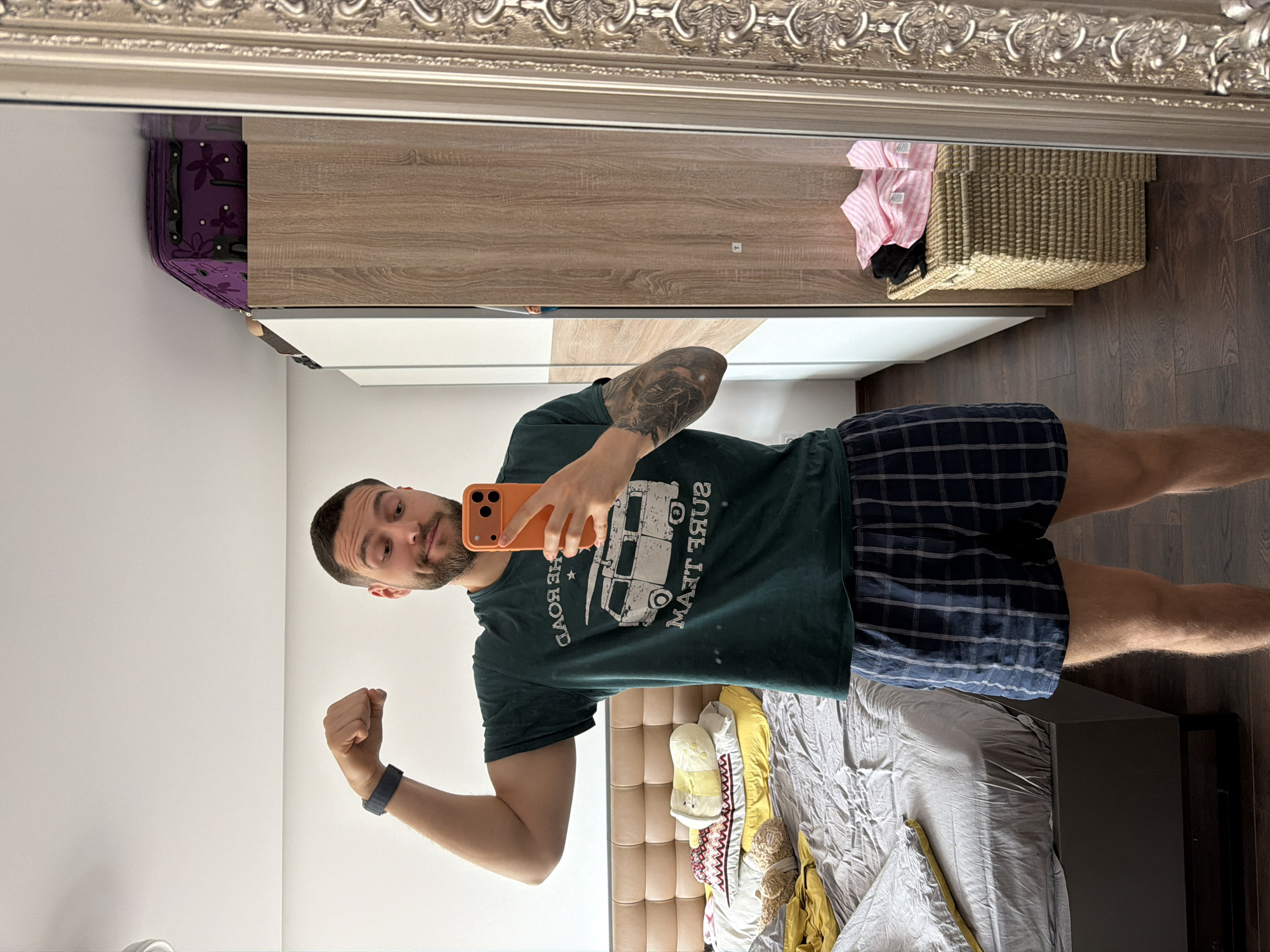 A man with short brown hair and a beard stands in front of a mirror in a room, taking a selfie with an orange smartphone. He is wearing a green T-shirt with a van design and plaid shorts. He flexes his left arm, displaying a dark-colored wristband.
The room contains a bed with a grey comforter and yellow, white, and red clothing items. A closet with white and light wood-grain doors stands behind him. Above the closet on the wall to the left is a purple bag. To the right of the closet is a wicker basket with pink and black clothing.
The frame of the mirror is ornate and silvery. The room appears to be located in an apartment building, as this is characteristic of Sofia, Bulgaria. There are no visible landmarks or information to confirm the exact address in Sofia.