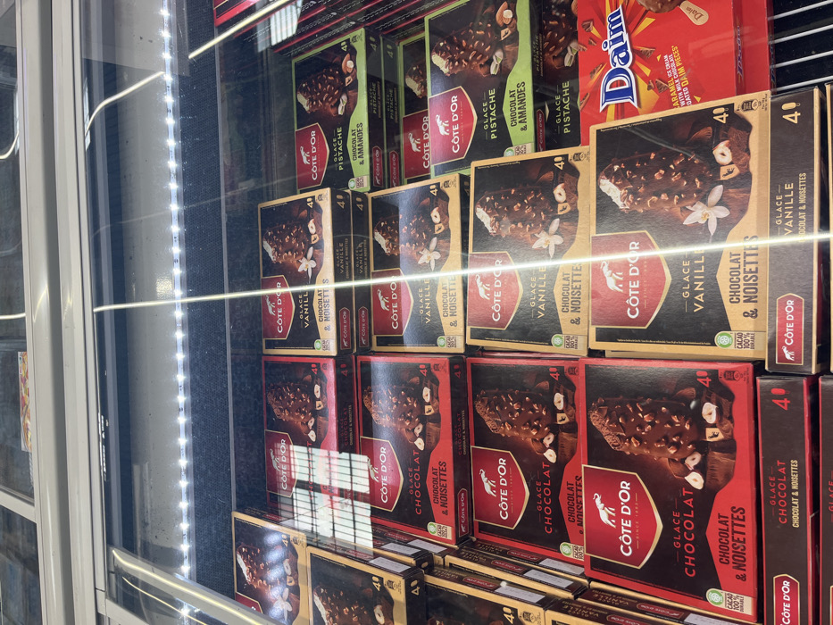 A refrigerated display case in a grocery store in Saint-Brice-Courcelles, France, stocked with boxes of "Côte d'Or" brand ice cream bars. The boxes are arranged in a grid pattern, each containing four ice cream bars. The ice cream flavors are chocolate, vanilla, and pistachio. The boxes all have the same design, with a gold background, a red or green band, and a picture of an elephant holding a scoop of ice cream. The price is 4.00 euros per box. The display case has an overhead light shining down on the ice cream. The display case is lit from above by fluorescent lights. The store's fluorescent lights are casting a warm glow on the scene. There is a box of "Daim" caramel ice cream to the right of the Côte d'Or ice cream bars. The Daim ice cream is packaged in a red and yellow box, with a picture of a caramel ice cream bar.