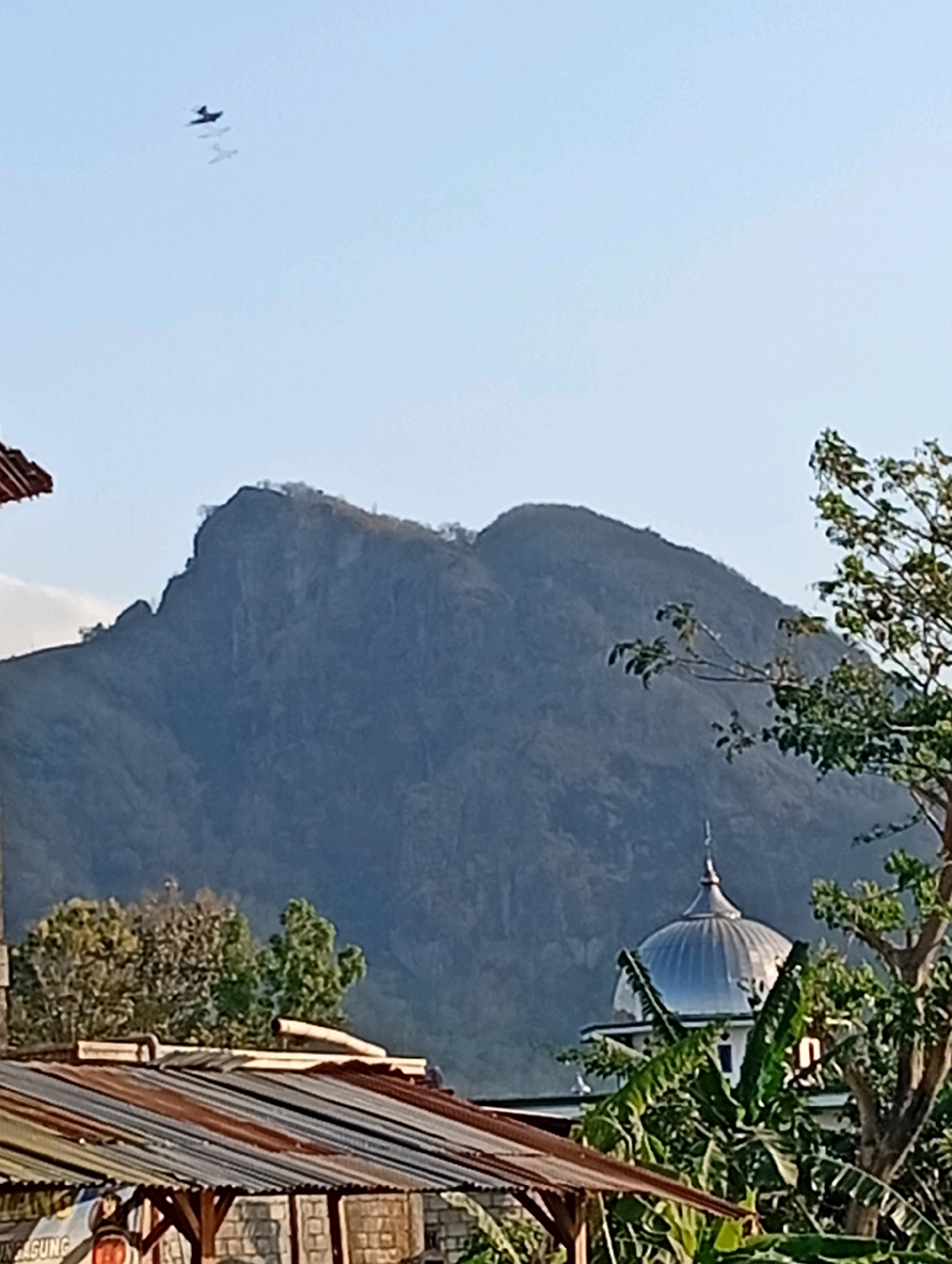 The image shows a view of a mountain in Sanggrahan, Indonesia. In the foreground, there are several rooftops, some made of corrugated metal, and some greenery including banana trees. Behind the rooftops, a domed building, likely a mosque, is visible. The mountain dominates the background, with a rugged, dark silhouette against a light blue sky. In the sky above the mountain, three small, dark shapes resembling airplanes are flying in formation. The overall scene appears to be a rural or semi-rural landscape.