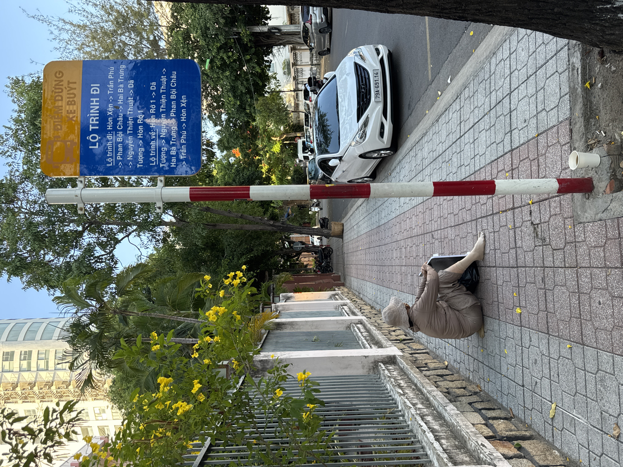 A person wearing a beige hat and matching clothing sits on a sidewalk in Nha Trang, Vietnam, facing away from the camera and towards a bus stop sign. The sign, a rectangular board with blue and yellow text, lists bus routes with destinations such as Hon Xen, Tran Phu, and Phan Boi Chau. Yellow flowers bloom on a tree to the left, casting dappled light on the scene. A white car is parked on the street next to the sidewalk. The sidewalk is paved with interlocking bricks.