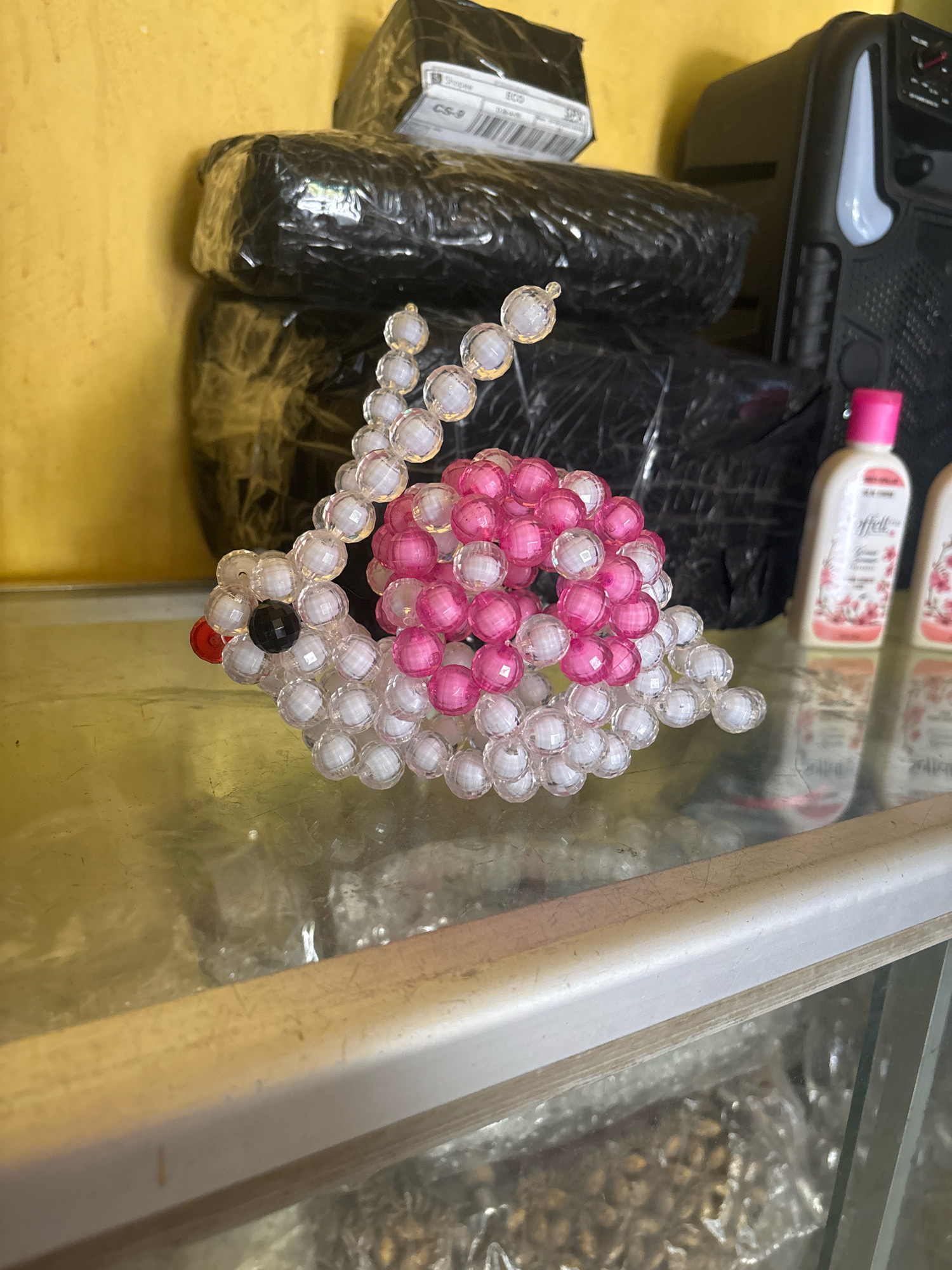 A beaded snail ornament, made of pink and clear beads, sits on a glass surface. Behind it are black plastic-wrapped packages and a portable speaker. In the background, a yellow wall is visible. To the right, two small bottles with pink caps and floral designs are present. The scene suggests an indoor setting, possibly a home or shop, in Jambi, Indonesia.
