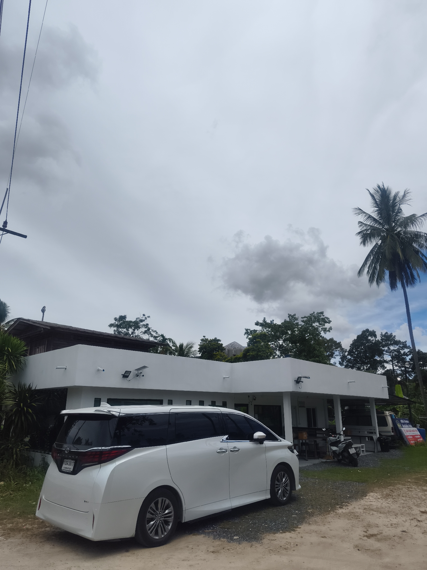 The image shows a white minivan parked on a gravel driveway in front of a white, modern building. The building appears to be a single-story structure with a flat roof and several windows. There is a palm tree to the right of the building, and other trees and vegetation are visible in the background. The sky is overcast with some clouds. The minivan is facing away from the camera, and its rear lights are illuminated. A license plate is visible on the back of the minivan. There are also a couple of motorcycles parked in front of the building. The scene suggests an outdoor setting, possibly a business or a residential area, in a tropical or subtropical climate.