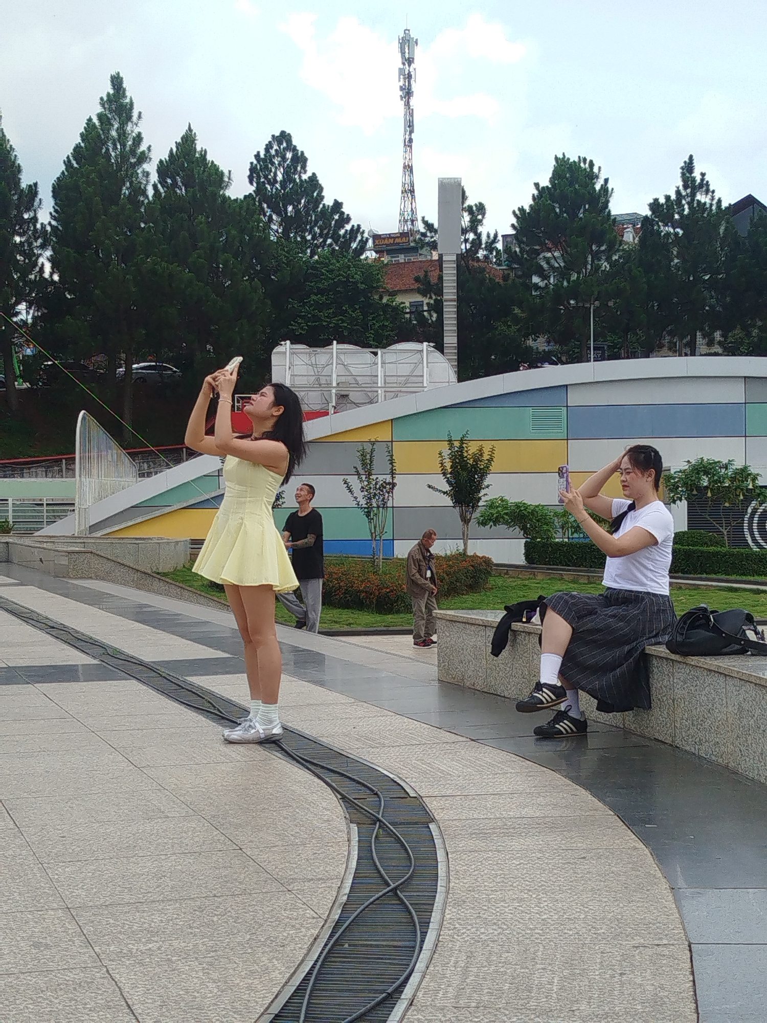 The image captures an outdoor scene in Da Lat, Vietnam, featuring a paved area with curved metallic channels. Two young women are present. On the left, a woman in a yellow dress stands and takes a picture with her phone. On the right, a woman in a white t-shirt and a grey skirt sits on a stone ledge, also engaged with her phone. In the background, other individuals are visible, along with trees, a colorful building facade, and a telecommunications tower. The sky is overcast.