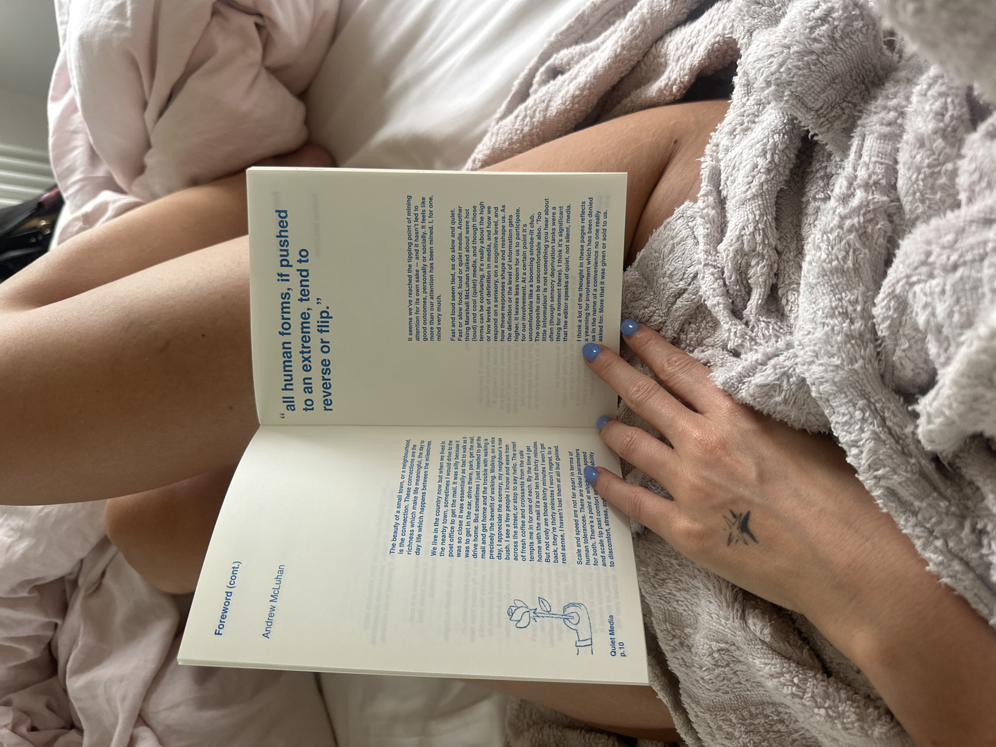 A person is lying in bed, holding open a book titled "Quiet Media." The book is open to a page that includes the quote "all human forms, if pushed to an extreme, tend to reverse or flip." The text on the opposite page includes "Foreword (cont.)" by Andrew McLuhan, along with "Quiet Media p.10." A visible tattoo is on the hand holding the right side of the book. The bedding is a mix of pink sheets and a gray textured blanket. The image appears to be taken indoors. There is no information available to establish the location in Tunbridge Wells, United Kingdom.