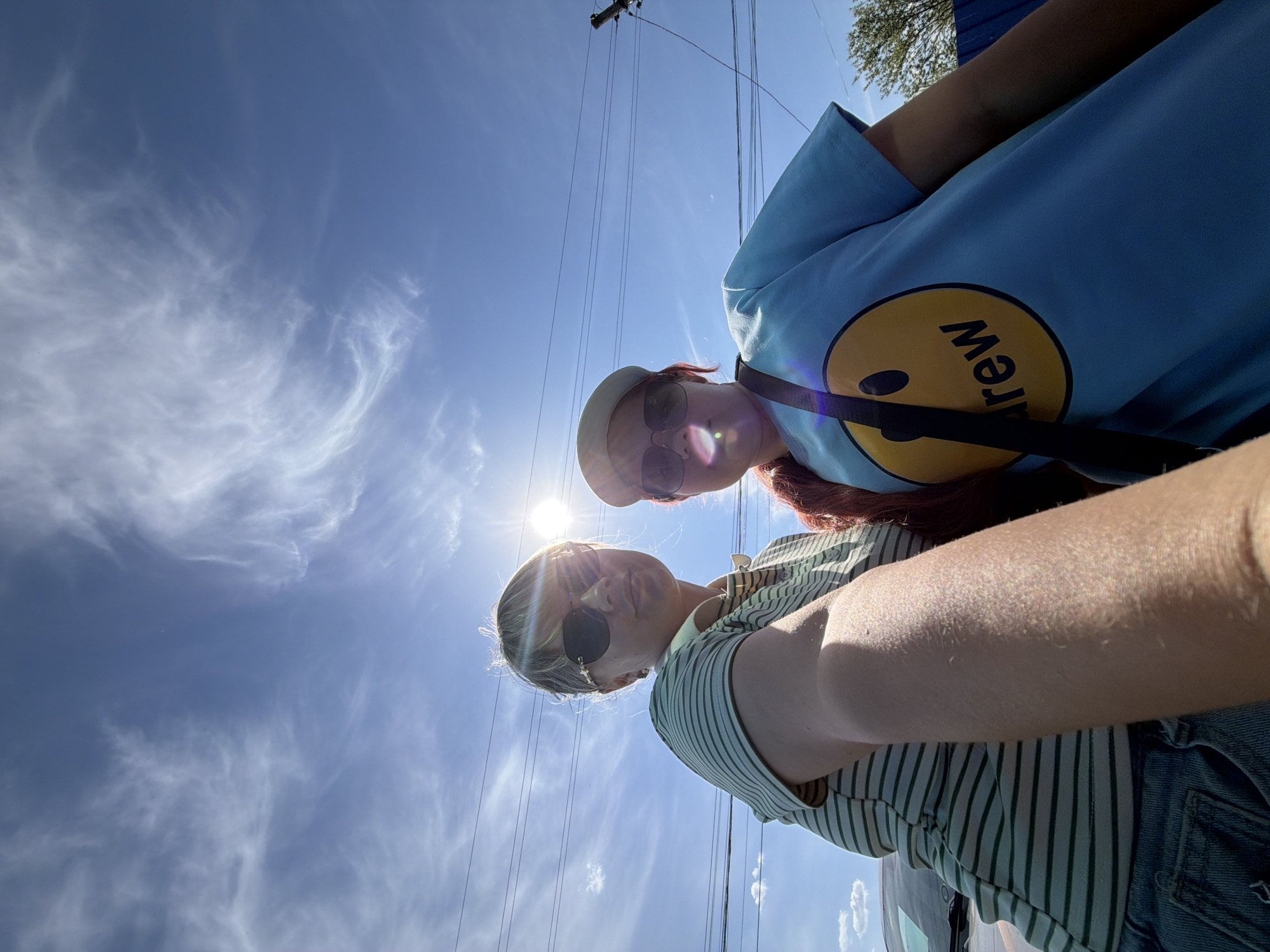 Two individuals, one wearing a light blue t-shirt with a yellow smiley face graphic and a white hat, and the other wearing a green and white striped shirt and sunglasses, are positioned close together, seemingly taking a selfie from a low angle looking upwards. The background includes a bright blue sky with wispy clouds, and overhead power lines are visible. The location cannot be determined from the image.