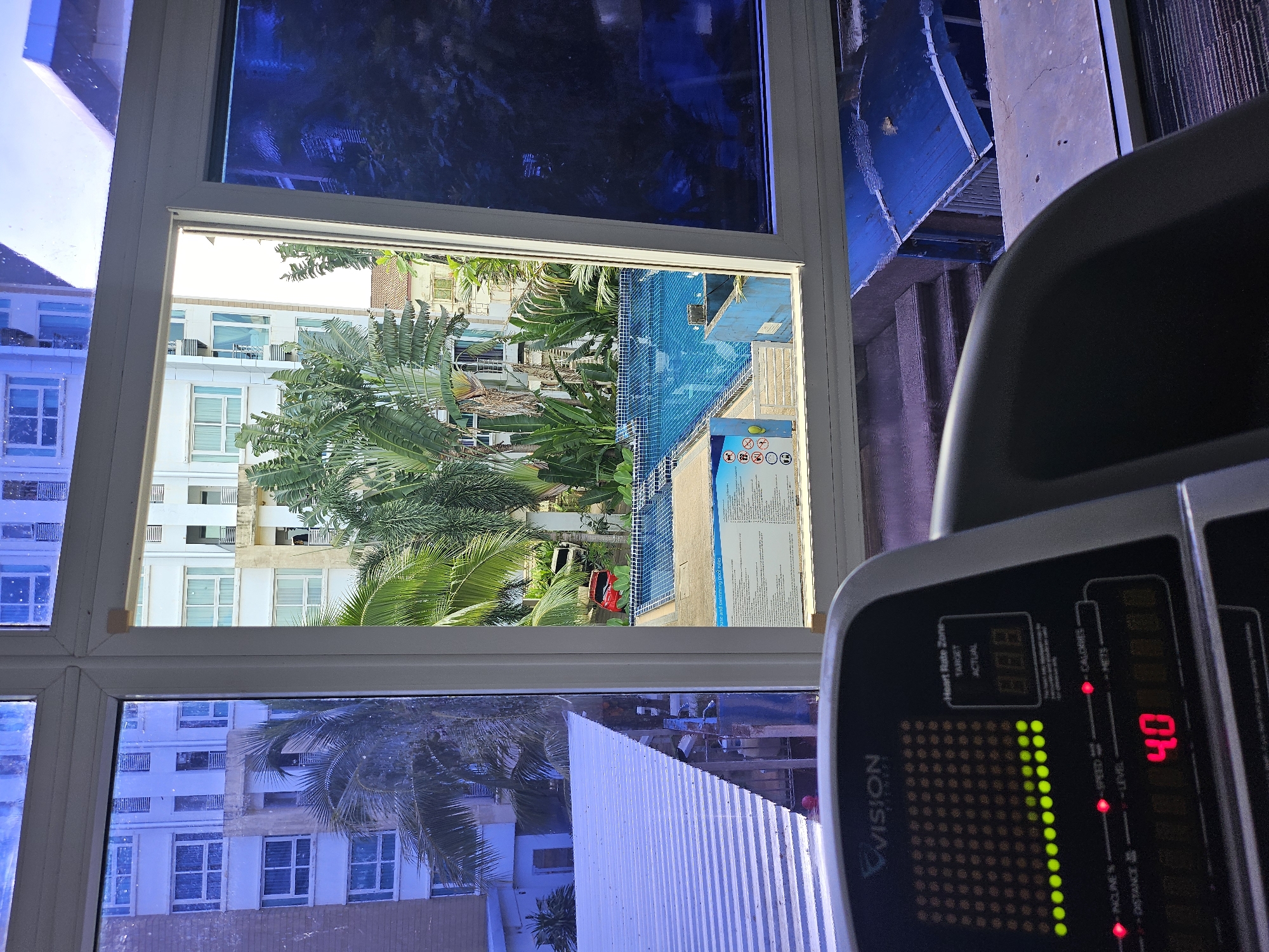 The image is taken from the perspective of someone inside a gym, looking out through a window. In the foreground, a treadmill console is visible, displaying "VISION FITNESS", and metrics such as "INCLINE %", "DISTANCE", "SPEED", "LEVEL", "CALORIES", and "METS". The speed is set to "4.0".
The window offers a view of an outdoor area with a swimming pool, surrounded by buildings and lush greenery, including palm trees. A red car is parked near the pool. A sign with swimming pool rules is also visible next to the pool. The buildings appear to be apartment complexes. The sky is bright, suggesting daytime.
The location is identified as Kathu, Thailand. The overall scene depicts a resort-like setting with indoor fitness facilities and an outdoor recreational area.