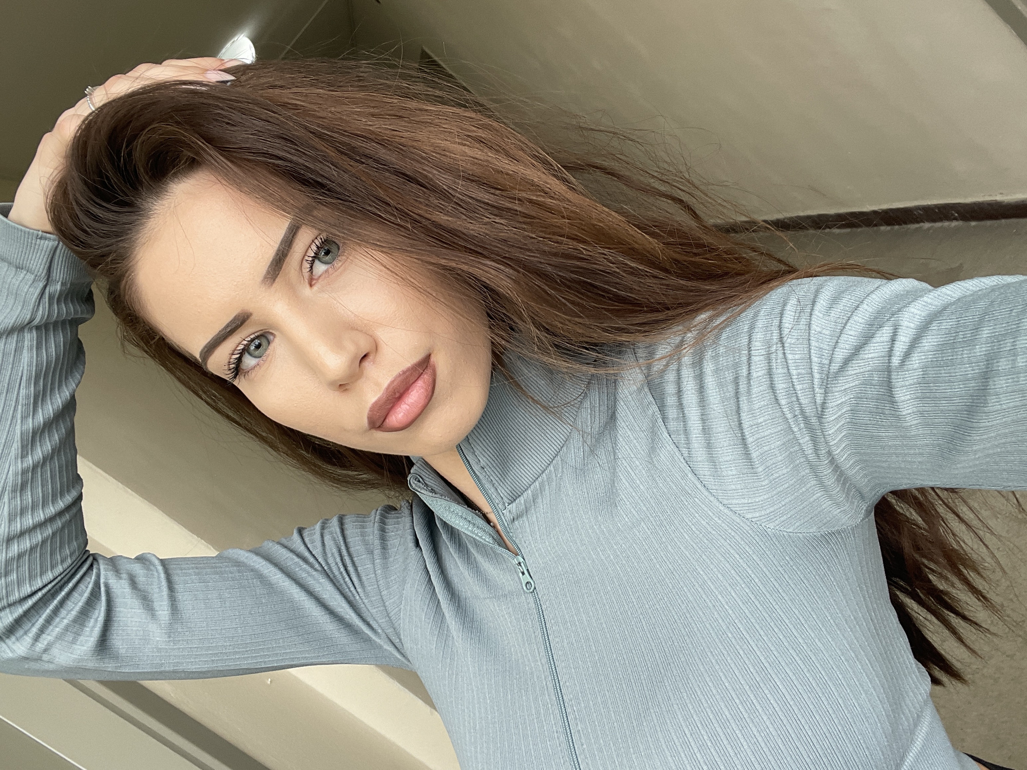 A young woman with long, auburn hair and light eyes is posing for a selfie. She is wearing a gray, ribbed, long-sleeved top with a zipper detail. Her right hand is raised, partially obscuring her hair. The background is a neutral-colored wall. There is no indication in the image of the city of Veliko Tarnovo, Bulgaria.