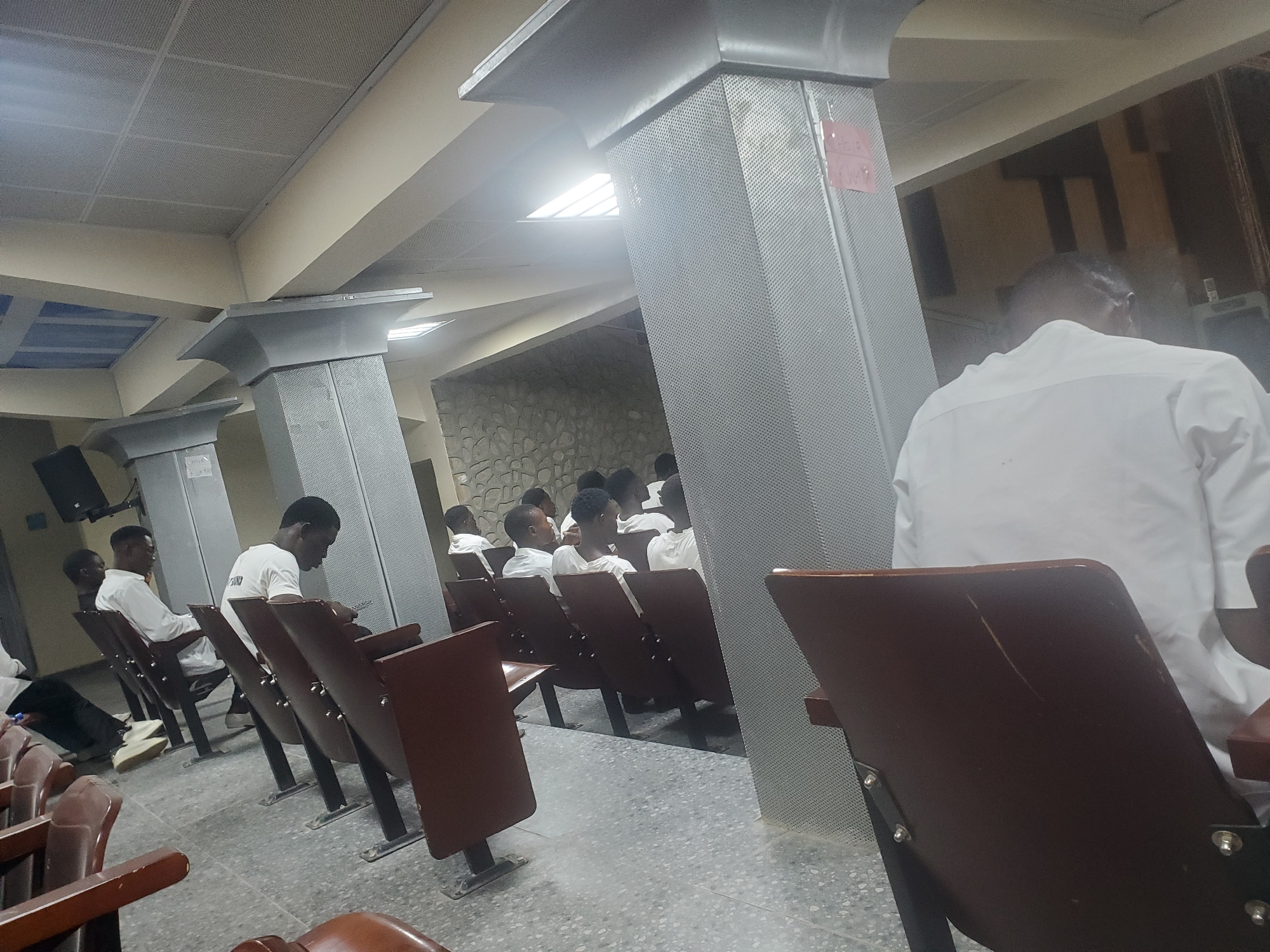 The image depicts an indoor scene, likely a hall or auditorium, in Kason, Nigeria. The environment features rows of dark brown chairs with metal legs, suggesting a seating area for an audience. Several individuals, predominantly young men wearing white shirts or garments, are seated in these chairs, facing towards a common direction, possibly a stage or presentation area that is not fully visible. The architecture includes large, gray, textured pillars and a suspended ceiling with a grid pattern. The lighting appears to be from overhead fixtures and natural light coming from windows or skylights, creating bright spots. The floor is a speckled gray tile. The overall atmosphere suggests a gathering or an event taking place in a formal setting.