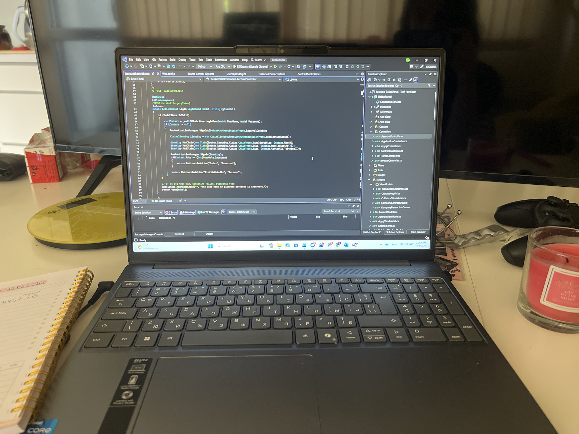 The image shows a laptop open on a desk, displaying lines of code in what appears to be a programming IDE. The laptop screen shows a file named "AccountController.cs" and other related files within a project named "BelizePortal". The keyboard of the laptop is visible, as is a portion of a notebook with handwritten text. To the right of the laptop, a glass candle with a pink hue is placed on the desk. The background shows parts of a room, including a television and some household items. The scene appears to be a home office or workspace. There are no people visible in the image. The metadata of the image indicates that it was taken on June 3, 2026, at 11:01 AM. The presence of Cyrillic characters on the keyboard suggests the user might be in a region where this alphabet is used, such as Bulgaria. However, there is no direct evidence in the image to confirm the location as Sofia, Bulgaria.