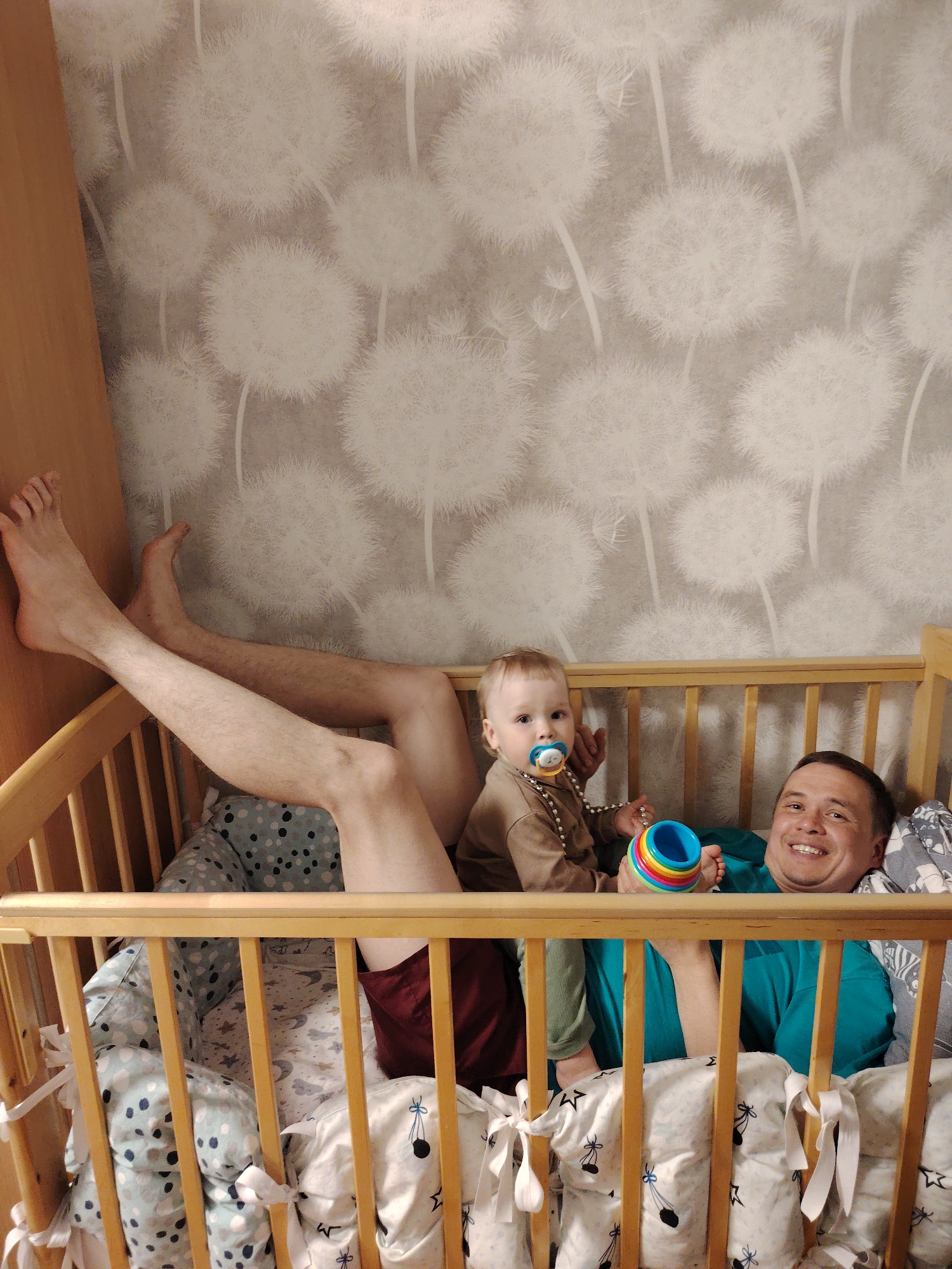 The image displays an indoor scene featuring a man and a baby, engaged in a playful moment within a wooden crib. The man is lying on his back, dressed in a teal t-shirt. His bare legs, wearing dark shorts, are extended upwards and rest against a light wooden piece of furniture on the left side of the frame. He is smiling broadly, looking directly towards the viewer.
Beside him, a baby or toddler sits upright. The baby is wearing a brown long-sleeved top, green trousers, and a beaded necklace. A pacifier is in the baby's mouth, and they are holding a colorful, ring-stacking toy. The baby also looks towards the camera with a neutral expression.
The crib is constructed from light-colored wood. The bedding includes padded bumpers with patterns of light blue and dark dots, and sheets featuring stars and small, stylized figures, which are secured to the crib bars with white bows. The background wall is covered in wallpaper depicting large white dandelion seed heads against a light grey background. The setting appears to be a child's room or bedroom, creating a soft atmosphere for this shared activity. No visible text is present in the image. The time of day is not discernible from the indoor lighting.