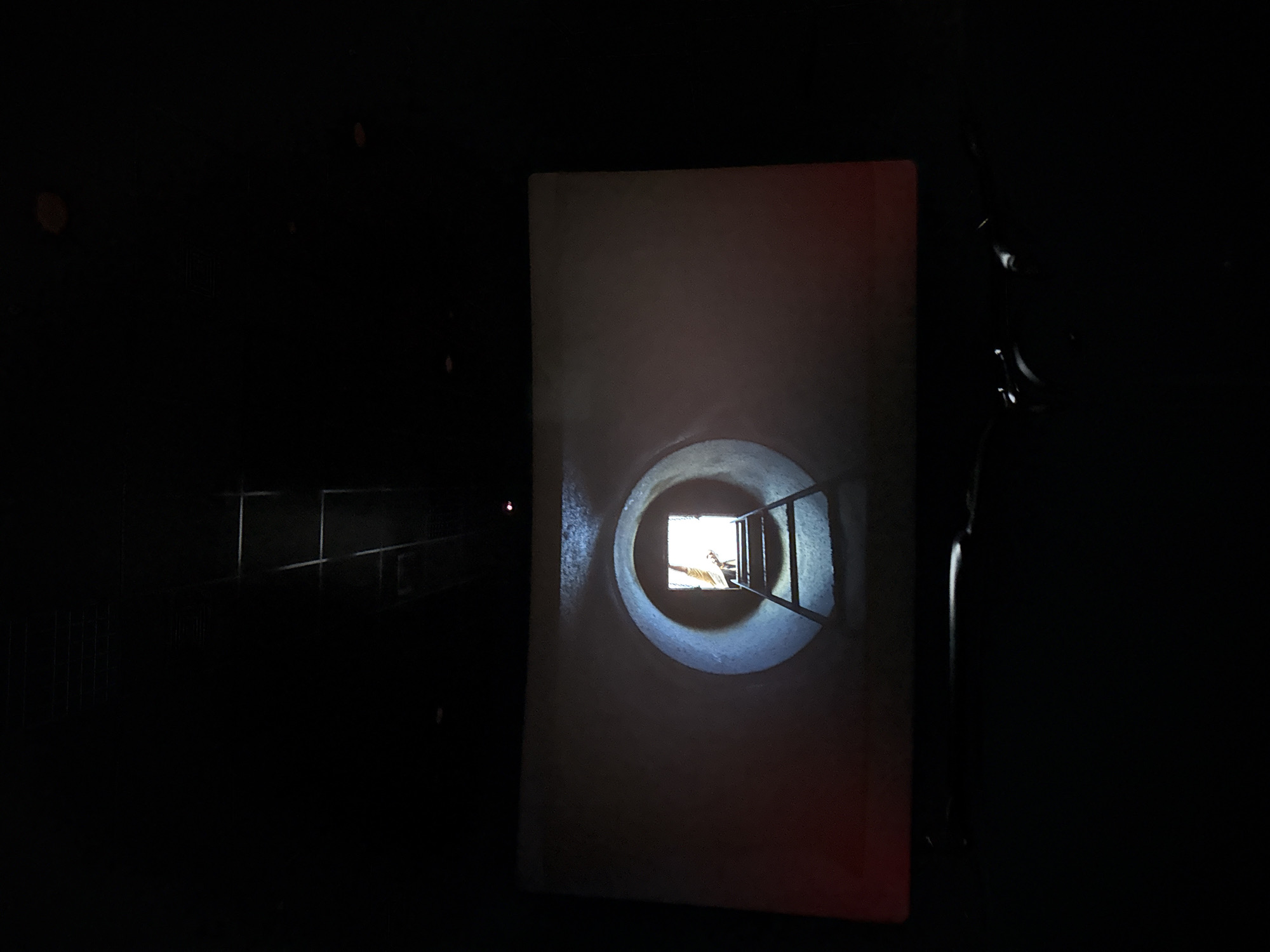 A dark, possibly indoor, scene features what appears to be a projected image of a round tunnel with a ladder leading to an exit at the end. The projected image is rectangular and centered in the frame. To the left, in the far background, faint outlines suggest some fencing or railing. To the right, indistinct shapes suggest a figure or object. The location and city are unknown.