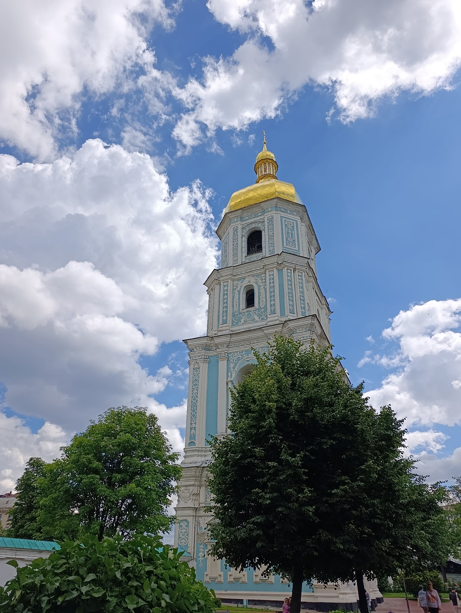The image shows a view of St. Sophia's Cathedral in Kyiv, Ukraine. The bell tower of the cathedral, with its golden dome, is the prominent feature. The tower is decorated with light blue and white ornamental details. Lush green trees are in the foreground, partially obscuring the lower part of the bell tower. The sky above is blue with fluffy white clouds. A few people can be seen in the background, walking along a path.