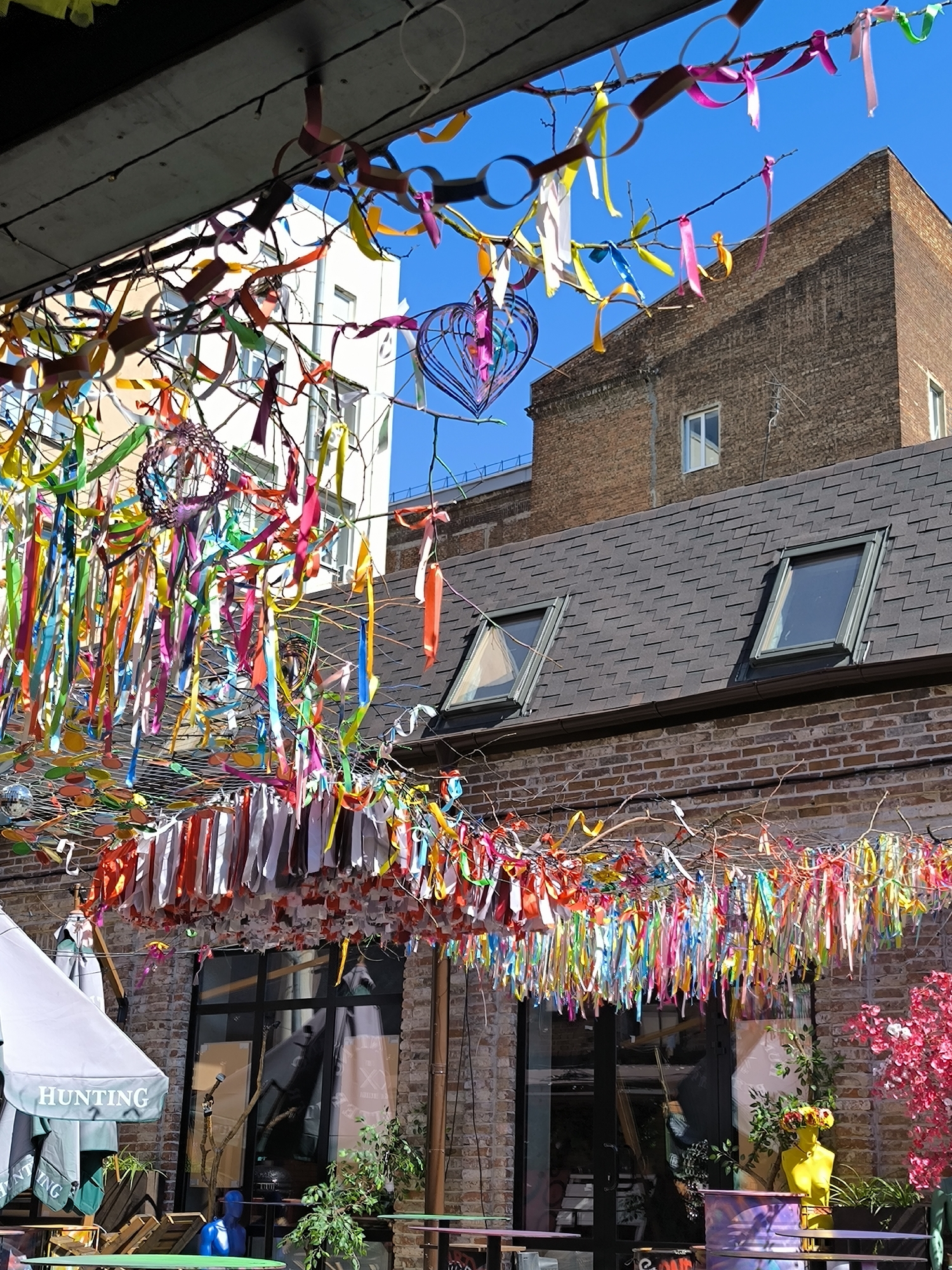 This media file shows an outdoor courtyard decorated with numerous colorful ribbons and streamers hanging from branches and wires. The ribbons are in a variety of colors including red, orange, yellow, green, blue, pink, and white, creating a festive and vibrant atmosphere. Some ribbons are tied in bows, while others hang loose.
In the foreground, there are outdoor tables and umbrellas. One umbrella is white and has the word "HUNTING" printed on it in a green font. There are also some plants visible in pots. In the background, there are brick buildings, one with a dark tiled roof and skylights. A bright blue sky is visible above. A large, intricate metal heart sculpture, also colorful, hangs among the ribbons. A yellow mannequin head with flowers on top is also present.
The scene appears to be set during the daytime, with bright sunlight casting shadows and illuminating the colors of the ribbons and decorations. The overall impression is one of a cheerful and celebratory outdoor space, possibly a cafe, restaurant, or event venue. There is no visible activity of people, but the decorations suggest an event or ongoing festive setup. The style of decoration evokes a sense of whimsy and joy.