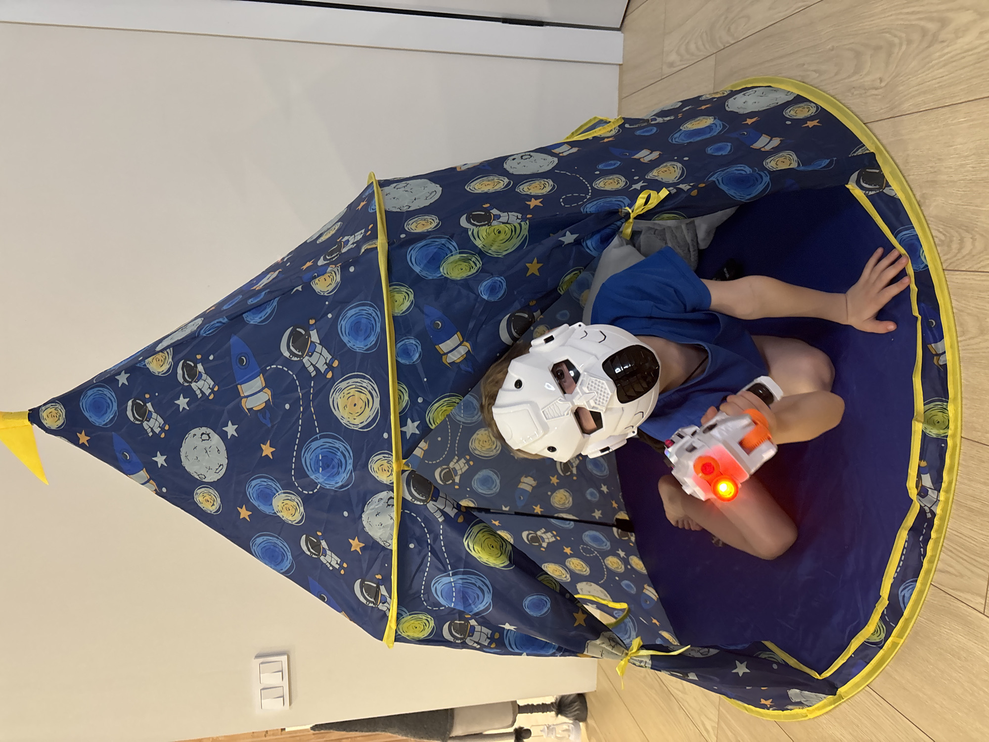 A child in a white helmet and blue shirt sits inside a blue play tent decorated with planets, stars, and rockets, holding a white and orange toy gun with a red light. The tent is indoors, placed on a wooden floor against a white wall. The location is the city of Chaikovsky, Russia.
