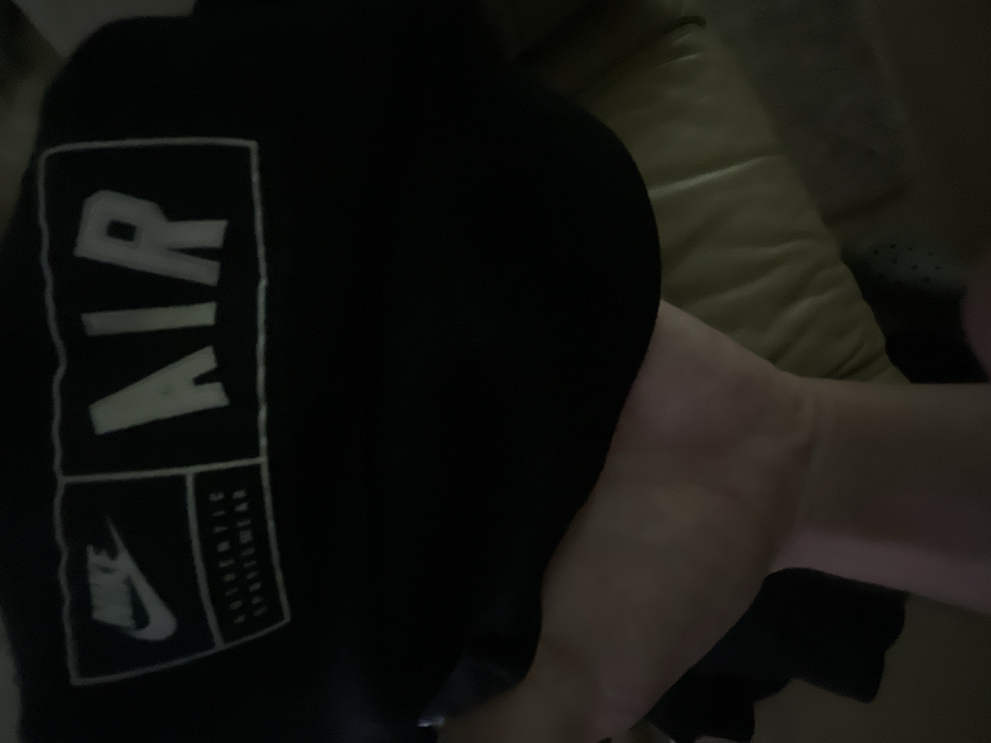 The image shows a close-up of a black t-shirt with a white "NIKE AIR" logo. The logo is in a rectangular box, with the Nike swoosh and the word "NIKE" on the left, and the word "AIR" on the right. Below the Nike logo, there is a smaller box with the words "AUTHENTIC SPORTSWEAR". A person's hand and arm are holding the t-shirt, obscuring part of the view. The background is dark and out of focus, but appears to be a textured surface, possibly a couch. There is no information in the image that can be used to determine its location in Bol'shoe Zaborod'e, Russia.