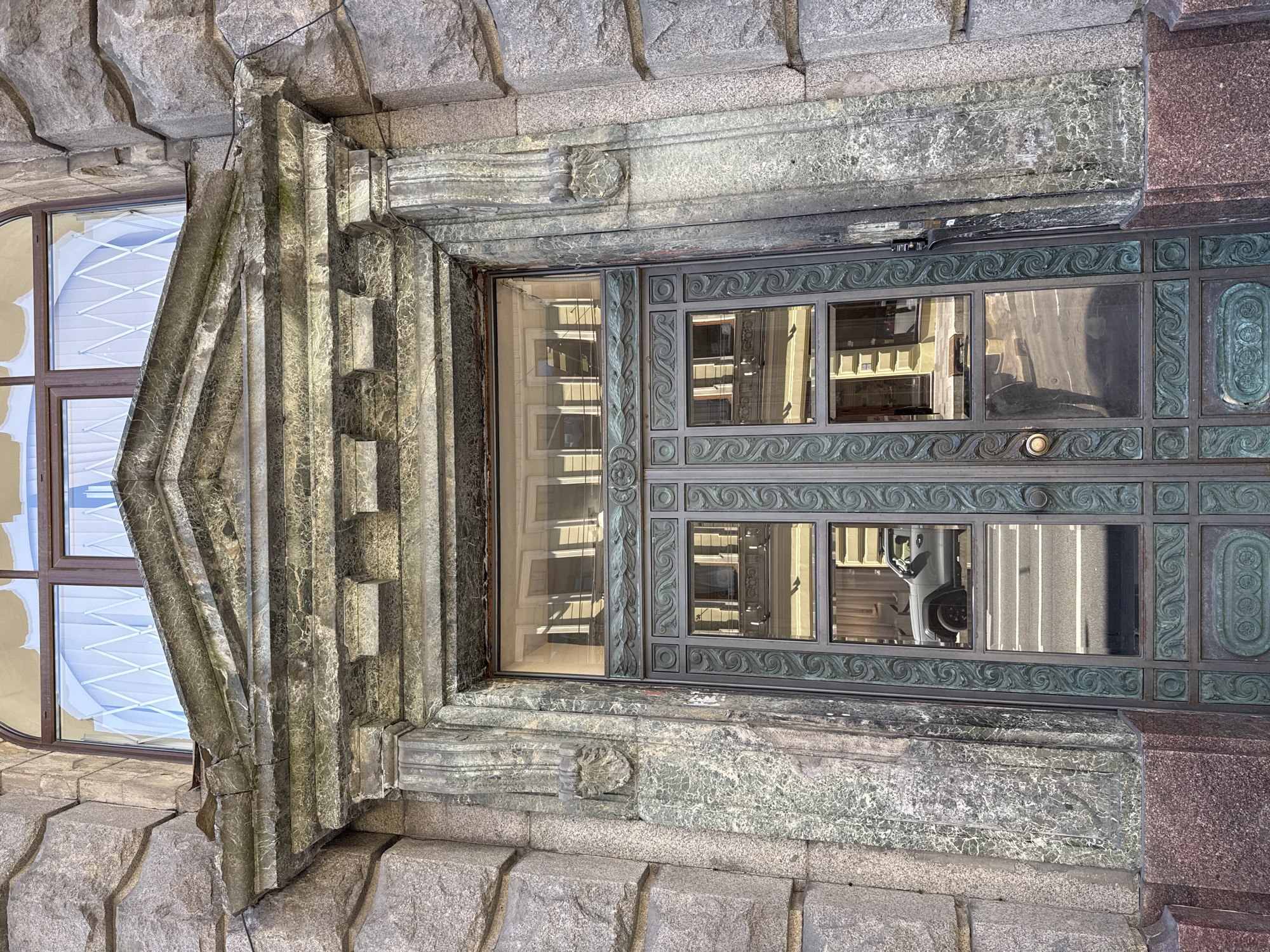 The image shows an ornate building facade with a large, detailed metal door and a window to its left. The door is dark green with intricate scrollwork and rectangular panels. One panel of the door reflects the opposite building, showing windows and a street. A round brass doorknob is visible. The window to the left of the door has a dark brown frame and a white crisscross pattern on the glass. The building's exterior is constructed from large, rough-hewn stone blocks at the base, transitioning to smoother, patterned stone and marble above. A carved stone archway, possibly part of an entrance, frames the door and window. The lighting suggests it is daytime, with shadows indicating a sunny or partly cloudy day. The architectural style appears to be classical or art nouveau, with a focus on elaborate detailing and heavy stonework. No people are visible in the frame.