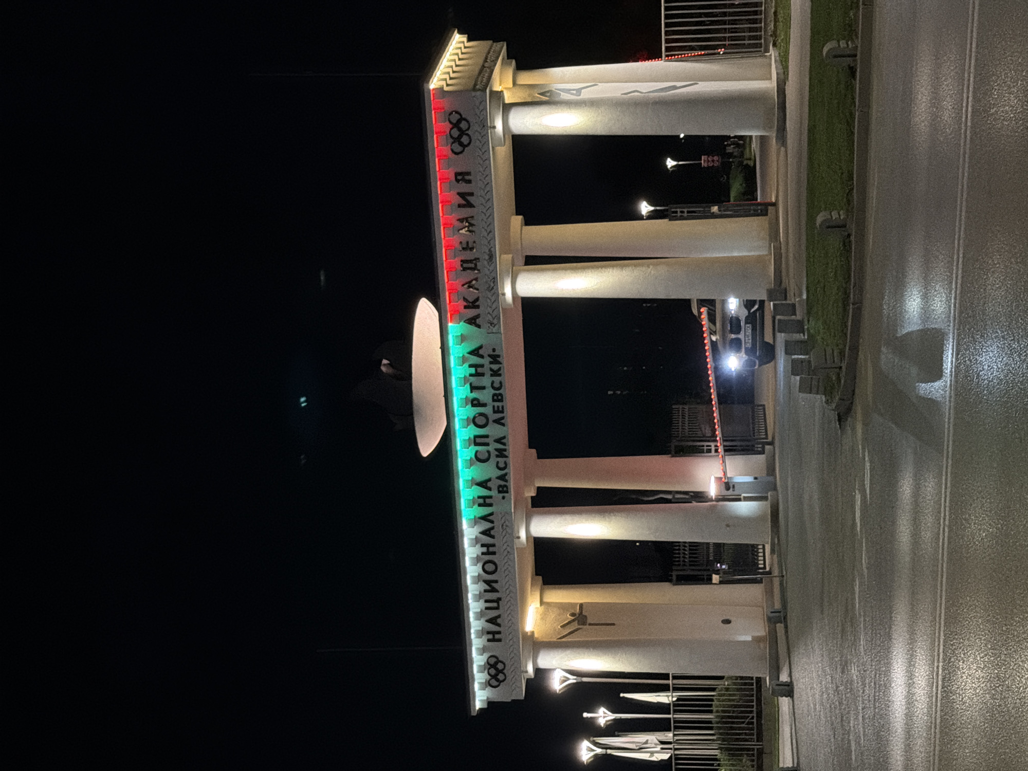 The image shows the entrance to the National Sports Academy "Vasil Levski" in Sofia, Bulgaria, at night. The entrance is a grand structure with large white pillars. A white banner above the pillars displays the name of the academy in Cyrillic letters, illuminated by red and green lights. The Olympic rings symbol is visible on either side of the banner. A car with its headlights on is approaching the entrance, which is secured by a barrier arm. The ground is wet, suggesting recent rain. Several lampposts with modern designs are also visible, along with some fencing and greenery. The overall scene is illuminated by artificial lights, creating a dramatic nighttime ambiance.