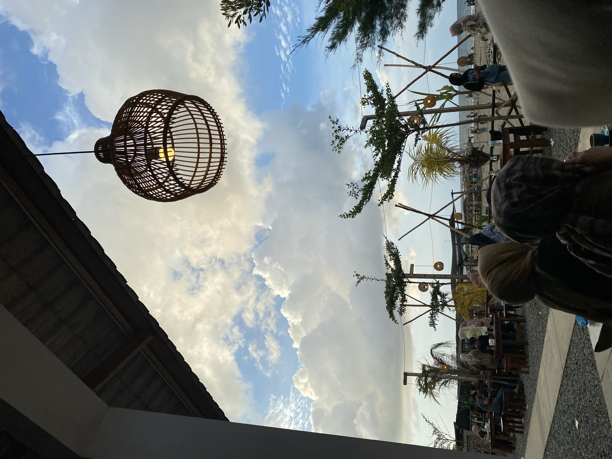 The image captures a scene in Keudekruenggeukueh, Indonesia, featuring an outdoor seating area likely part of a cafe or restaurant. The dominant feature is a large, spherical, wicker-like lamp hanging from the building's roof on the left, emitting a warm light. Above, the sky is filled with a mix of blue patches and voluminous, white and gray clouds.
The outdoor area is populated with wooden tables and chairs, occupied by several people, some partially obscured, suggesting they are patrons. Structures made of bamboo poles with attached decorative lamps are interspersed among the tables, adding to the ambiance. In the distance, the beach is visible, with more people along the shoreline. The overall environment suggests a relaxed, beachside dining setting.