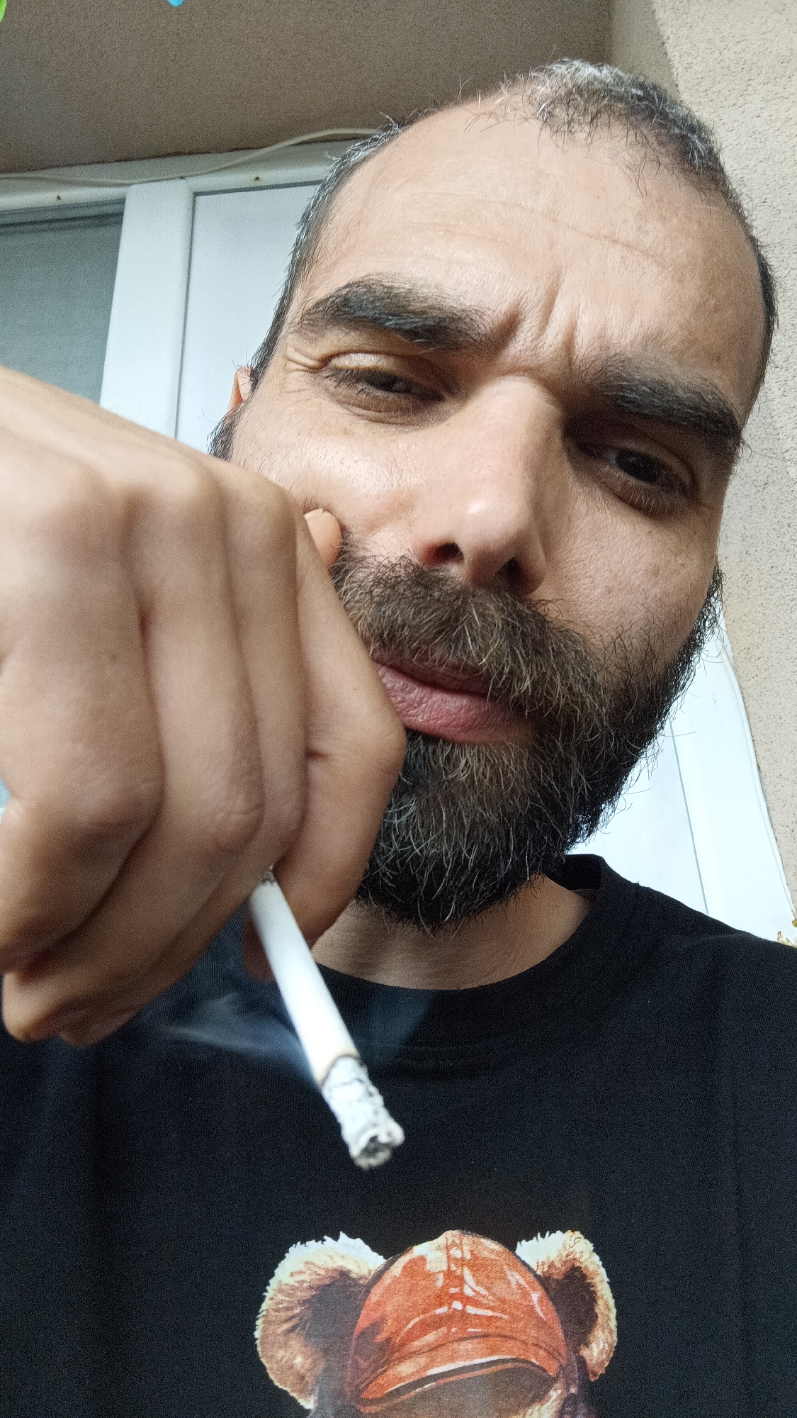 A man with a beard and grey hair is holding a lit cigarette. He is wearing a black shirt with an image of a bear. The background appears to be an exterior wall and a white door. Location: Unknown, Unknown.