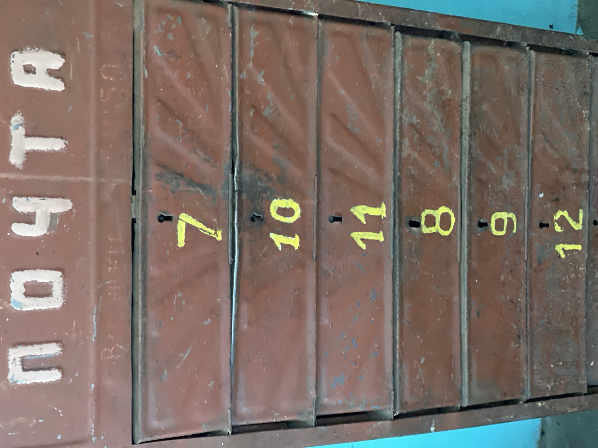 A close-up, eye-level, vertical shot shows a weathered, dark red, multi-drawer mailbox. The top of the mailbox has Cyrillic lettering in white with blue outlines that reads "ПОЧТА" (POCHTA), meaning "POST". Below this, there are faint, unreadable markings. Each of the six visible drawers has a yellow number painted on its front. From top to bottom, the numbers are 7, 10, 11, 8, 9, and 12. Small keyholes are visible above the numbers 7, 10, 11, 8, 9, and 12, suggesting each drawer is lockable. The metal of the mailbox is scratched and worn, with some areas showing rust. The background to the left of the mailbox is a bright turquoise wall, and to the right is a darker, possibly metallic, structure.