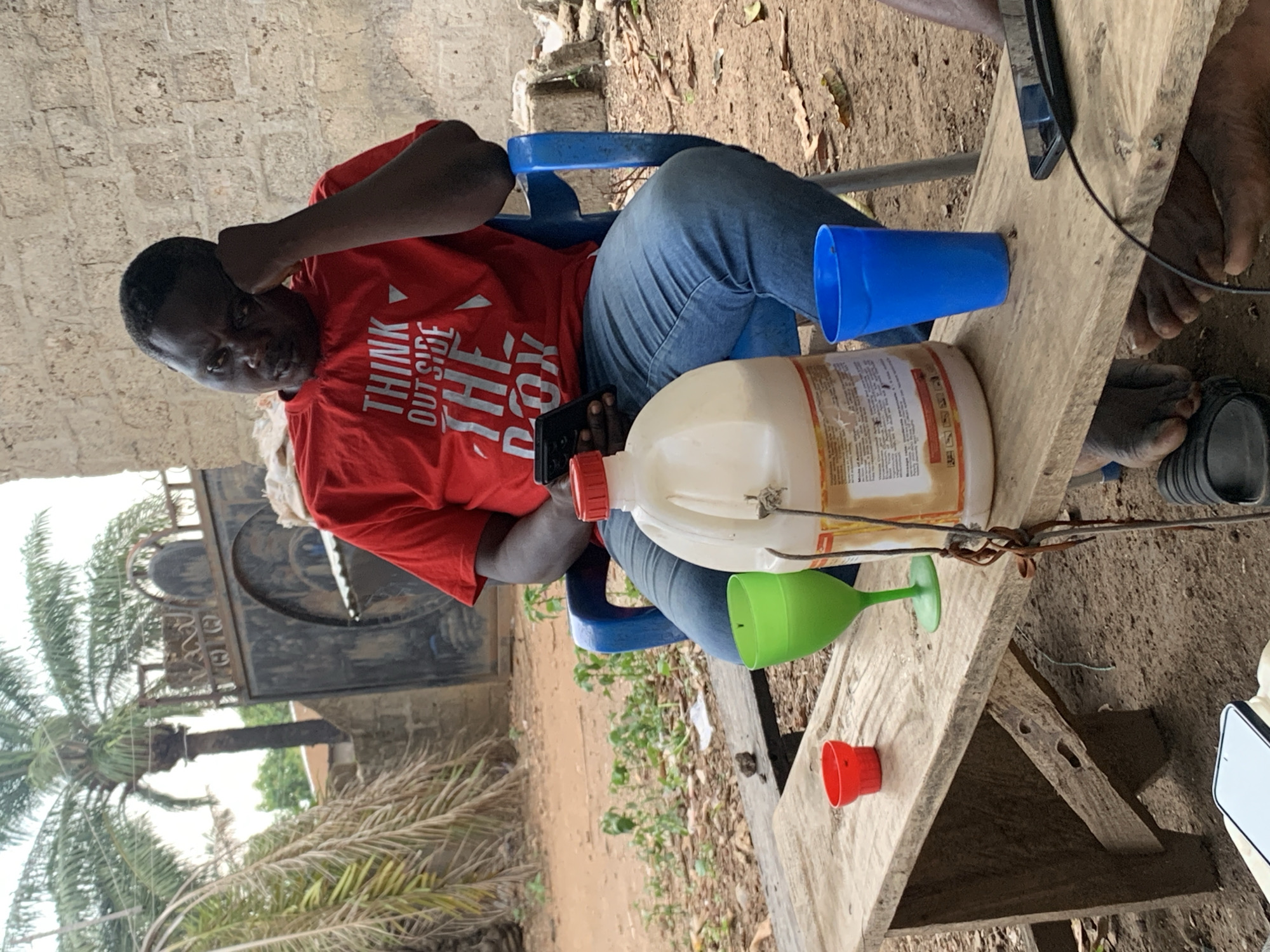 The image depicts a man sitting on a blue plastic chair, wearing a red t-shirt and blue jeans. He is holding a cell phone in his left hand and his right fist is pressed against his forehead. The man is sitting at a rough, wooden table. On the table are a blue plastic cup, a green plastic cup/funnel, a red plastic cup, and a large plastic container. An additional person's feet are visible in the upper right corner. The background shows a partially visible gate, palm trees and some foliage. The image was taken in Akwanga, Nigeria.