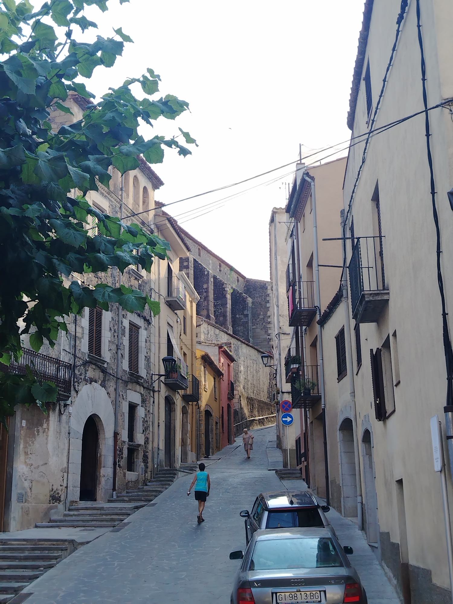 A narrow, uphill cobblestone street in Hostalric, Spain, is flanked by old buildings with textured facades and balconies. Green foliage from a large tree overhangs the left side of the image. Two individuals are visible: a person in a blue tank top and black shorts walks away from the camera up the street, and another person in a light-colored dress walks towards the camera in the distance. Two cars are parked on the right side of the street. A blue and white directional sign is visible on the right side of the street. The sky is bright and overcast.