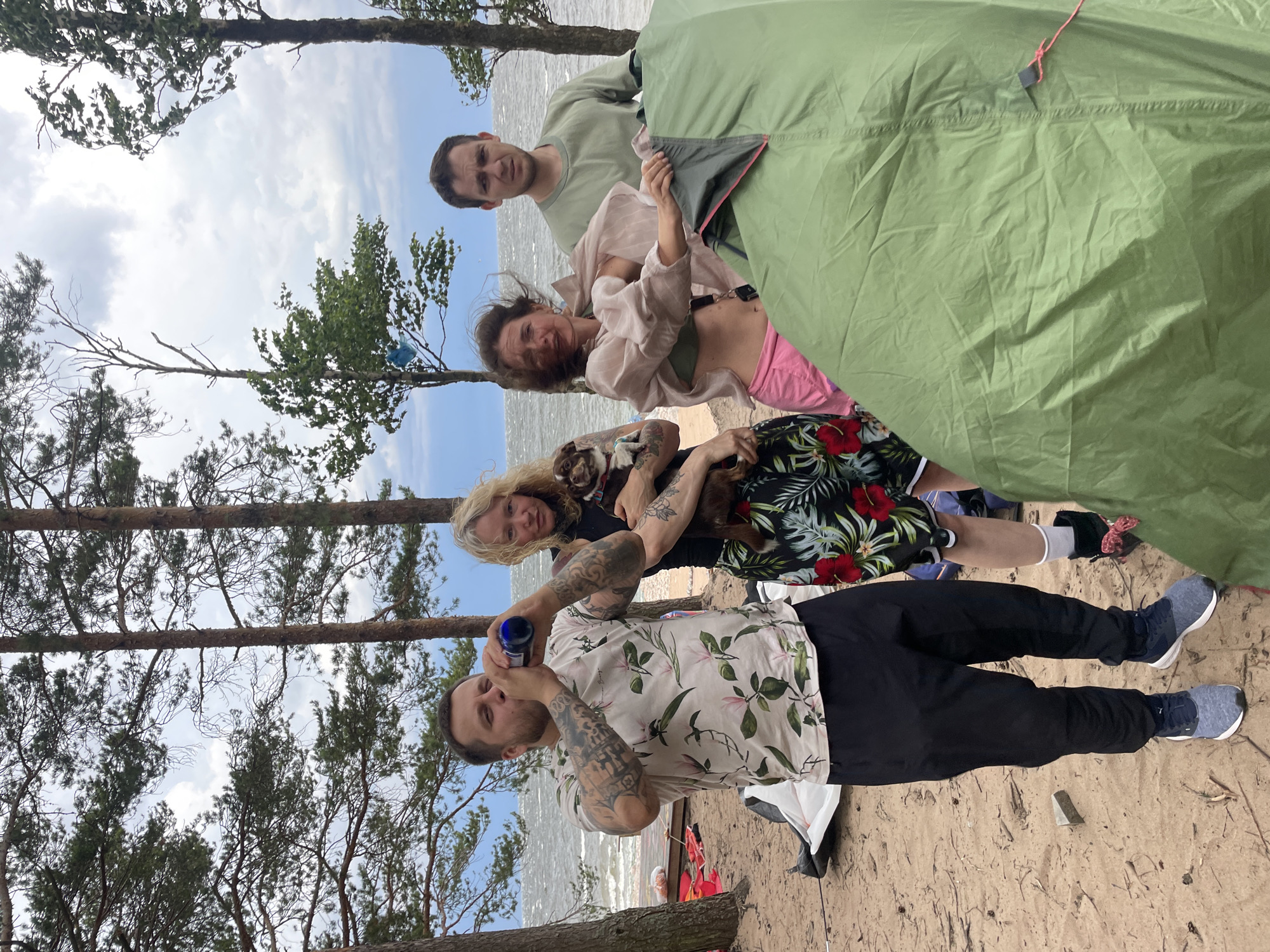 In Luzhki, Russia, on a sandy beach with a backdrop of the sea and overcast sky, four individuals and a dog are gathered around a partially set up green tent. The man on the left, wearing a floral short-sleeved shirt and black pants, is drinking from a blue bottle. To his right, a woman with blonde hair, in a black top and a floral skirt, holds a small brown dog. Next to her, a woman in a light pink sheer blouse over a green bikini top and pink shorts is in motion, possibly adjusting the tent, with her hair blowing in the wind. The fourth individual, a man in a green t-shirt and dark pants, stands to the right of the women, looking towards the viewer with a concerned expression. The scene suggests a casual outdoor gathering or camping trip.