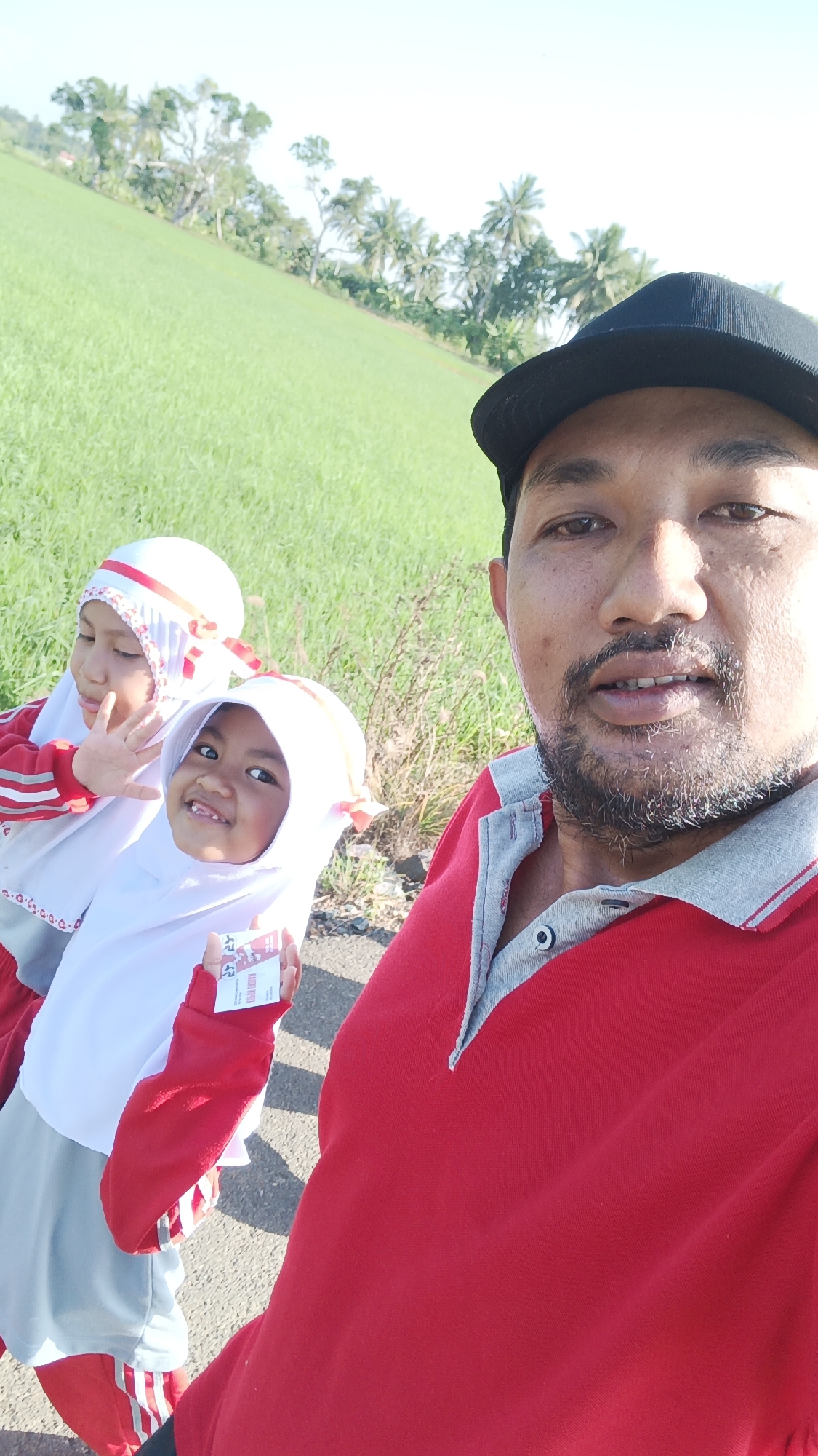 In Batarang, Indonesia, a man wearing a red polo shirt and a black baseball cap takes a selfie with two young girls. The girls are also dressed in red and white. One of the girls is waving at the camera. In the background, a lush green field stretches out under a bright sky, dotted with palm trees. The scene appears to be a casual outdoor setting, possibly a park or a rural area.