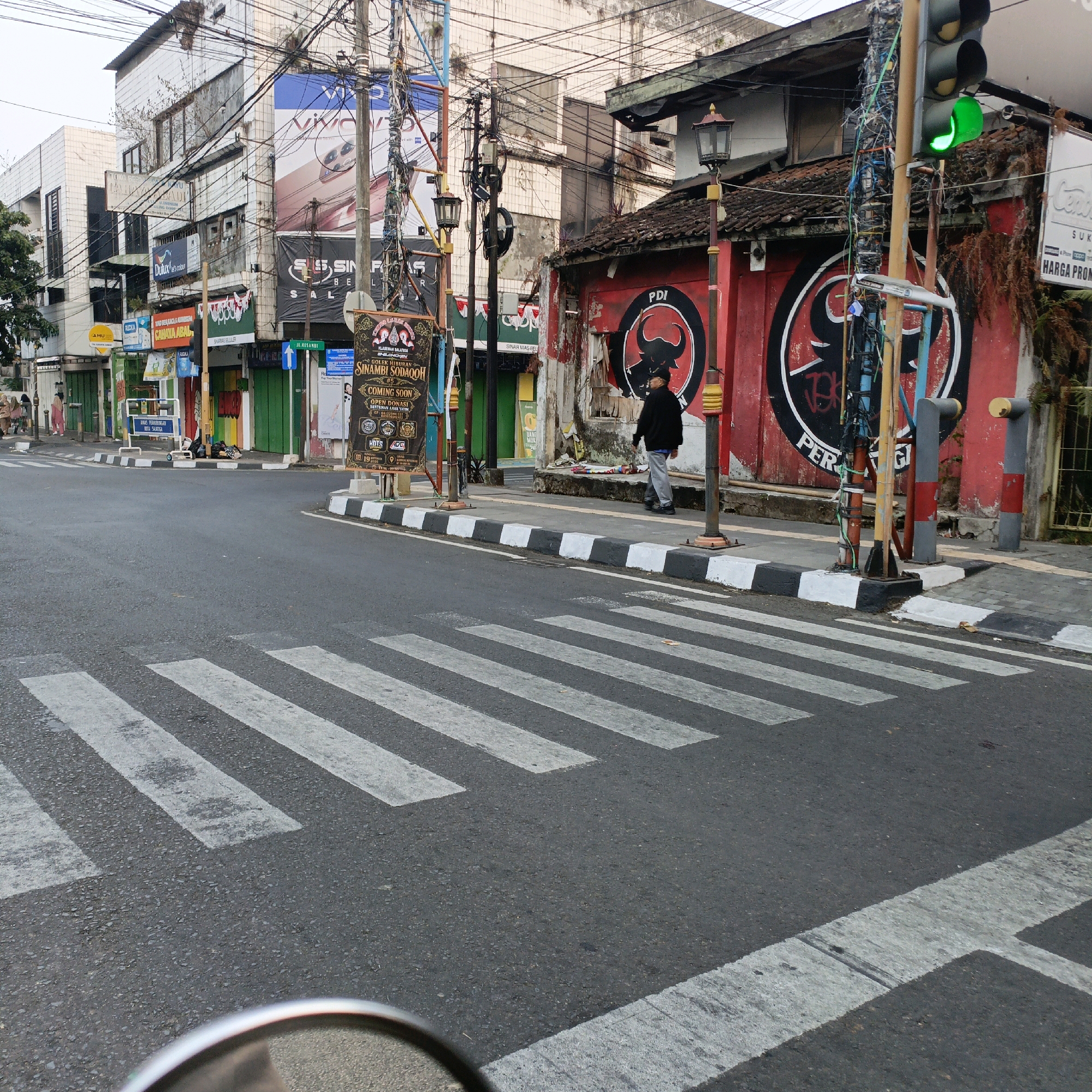 The image shows a street scene in Salatiga, Indonesia, with a crosswalk in the foreground and buildings on either side of the street. A traffic light is visible, showing a green signal. A lone person in dark clothing is walking on the sidewalk. The buildings appear to be commercial establishments, with some showing signs of age and disrepair. There are numerous utility poles and wires overhead, typical of urban infrastructure. The overall atmosphere suggests a somewhat quiet street, possibly early in the morning or during a less busy time of day. The presence of a crosswalk and traffic signals indicates it is an intersection within the city.