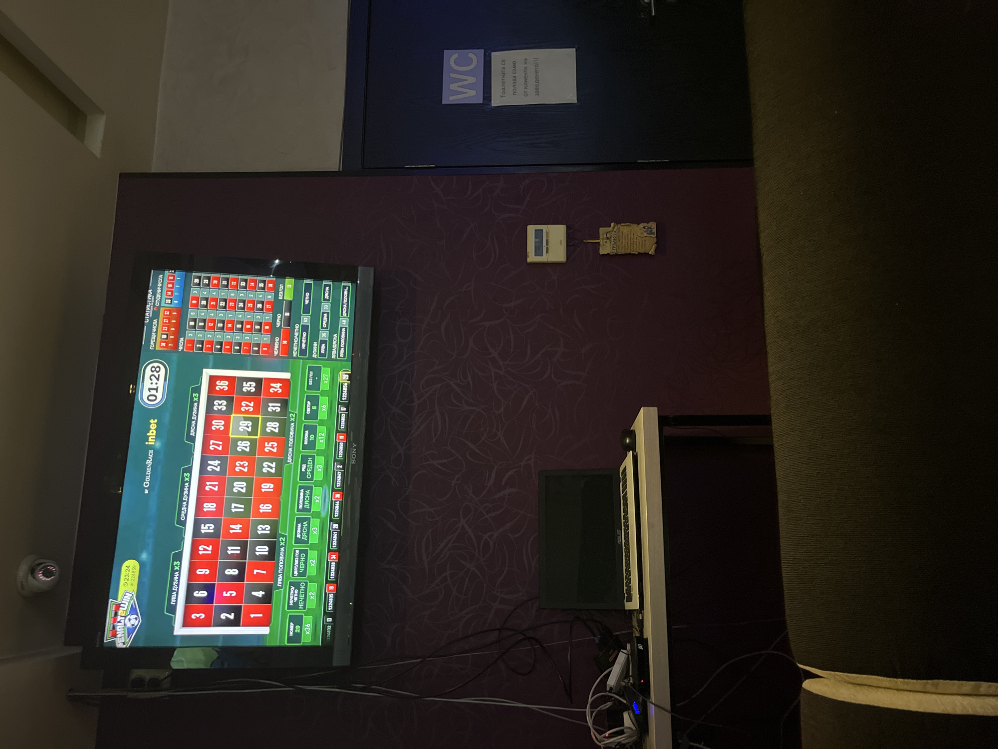 The image shows a television screen displaying a roulette game in a dimly lit room. The screen shows a betting layout with numbers and betting options such as "LEFT HALF X2", "RED", "BLACK", "ODD", "EVEN", etc. A timer on the screen shows "01:28". There are also statistics displayed on the screen, including a list of numbers and their frequency.
In the foreground, there is a dark-colored couch. To the right of the couch, a dark blue door is visible with a "WC" sign and a notice in Bulgarian that translates to "The toilet is for clients of the establishment only!!!". To the left of the door, a wall-mounted device is visible, possibly a thermostat or a control panel.
Further to the left, a desk or table is present, on which a laptop, a mouse, and some electronic devices are placed. Wires are running from these devices and connect to the wall. A security camera is mounted on the ceiling in the upper left corner of the image. The walls are decorated with a dark purple textured wallpaper.