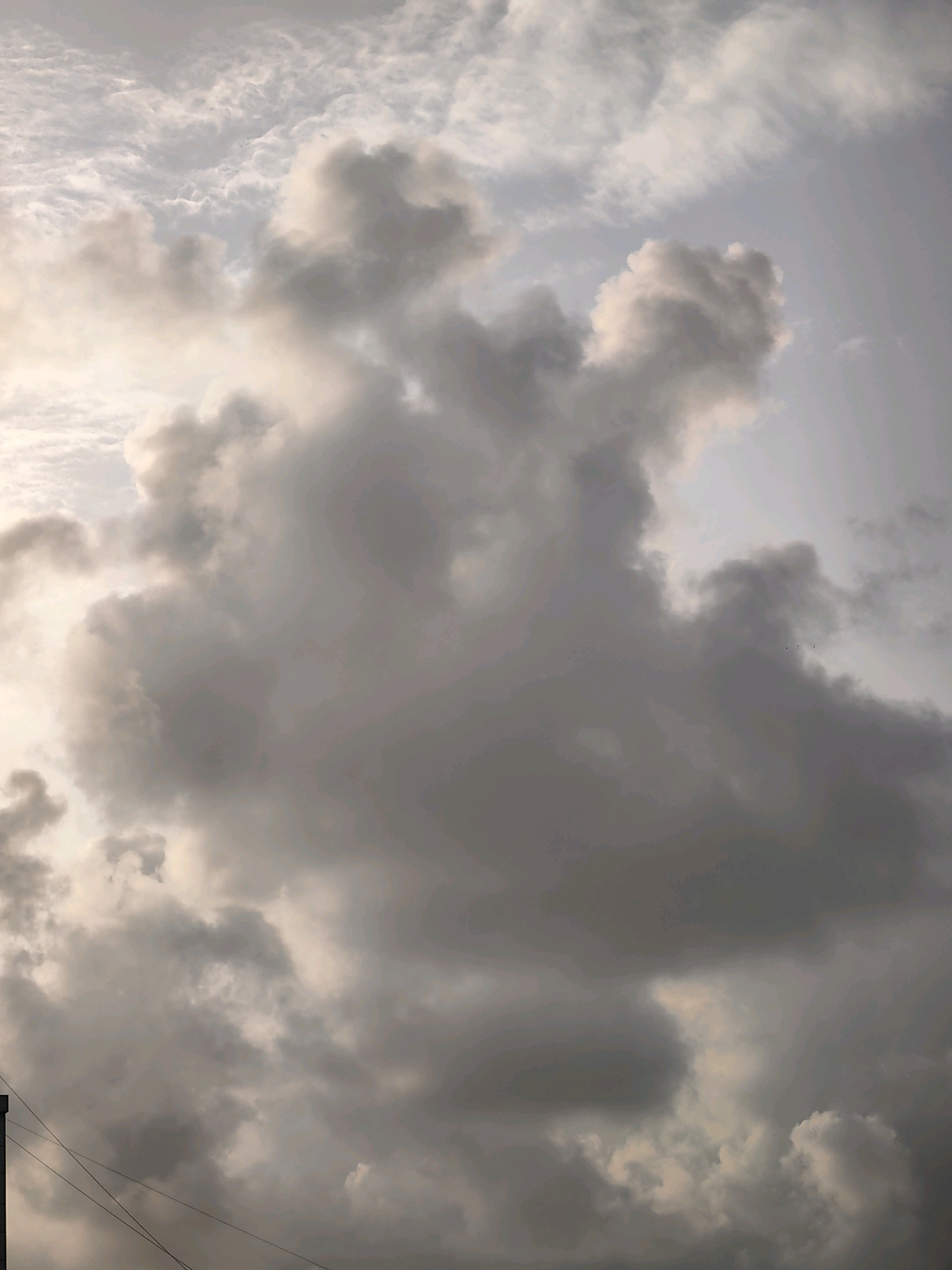 A vertical, eye-level medium shot captures a dramatic sky filled with dark, turbulent storm clouds. The clouds are densely packed and obscure most of the sky, with pockets of lighter, diffused light breaking through in the upper left. The overall mood is ominous and charged with the potential for rain. The lower portion of the image shows a sliver of the city of Mumbai, India, with the dark, vertical edge of a building on the far left, and a few thin, black electrical wires stretching horizontally across the bottom. There are no people visible in the frame.