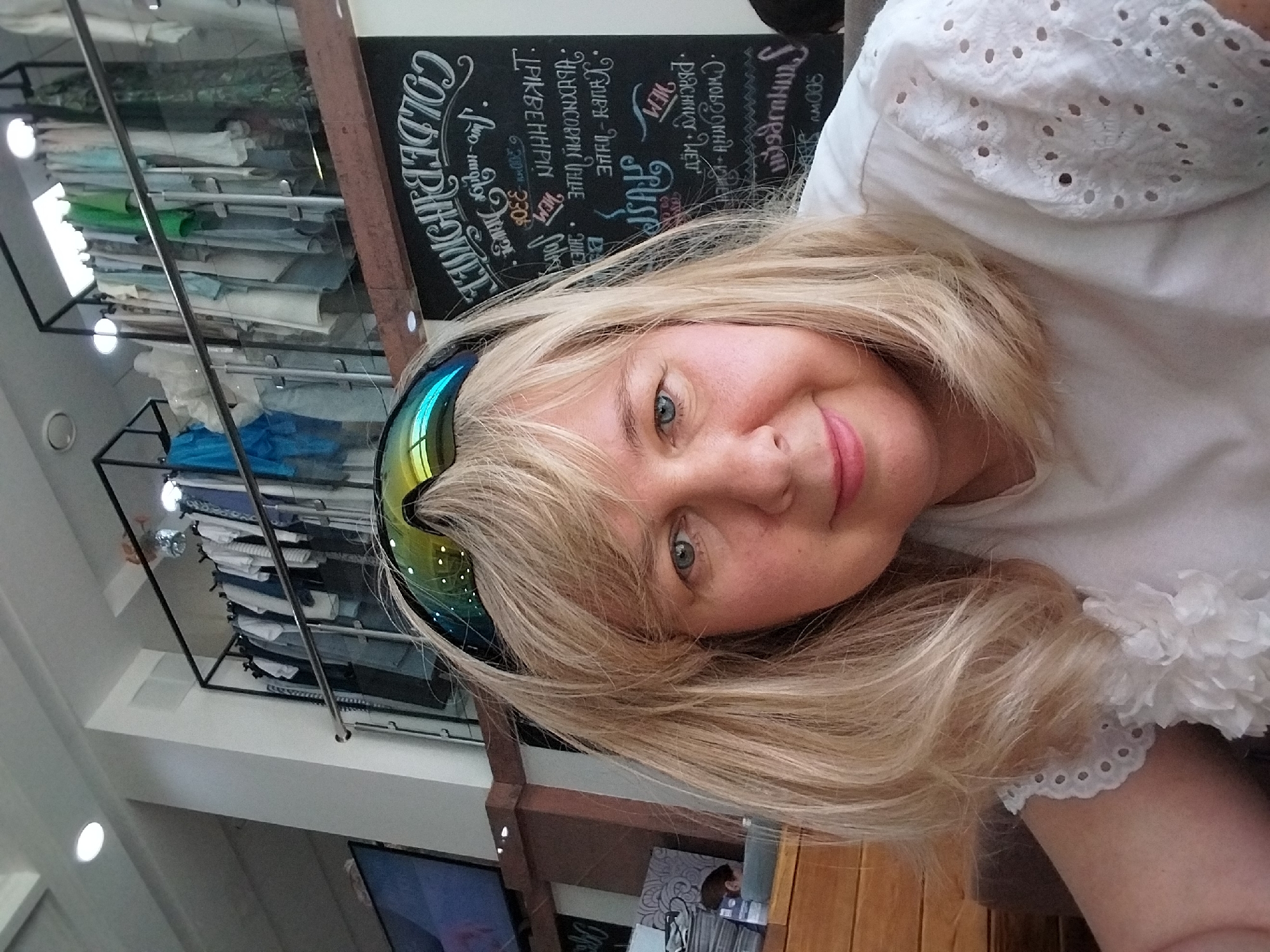 The image shows a woman with blonde hair and blue eyes wearing a white shirt with eyelet sleeves. She is wearing sunglasses on her head. Behind her, there is a rack of clothes hanging on a shelf, and a blackboard with writing on it. A television screen is visible on the left side of the image. The overall scene appears to be indoors, possibly in a retail establishment or cafe. The context of Orenburg, Russia, is not evident from the visual information provided.