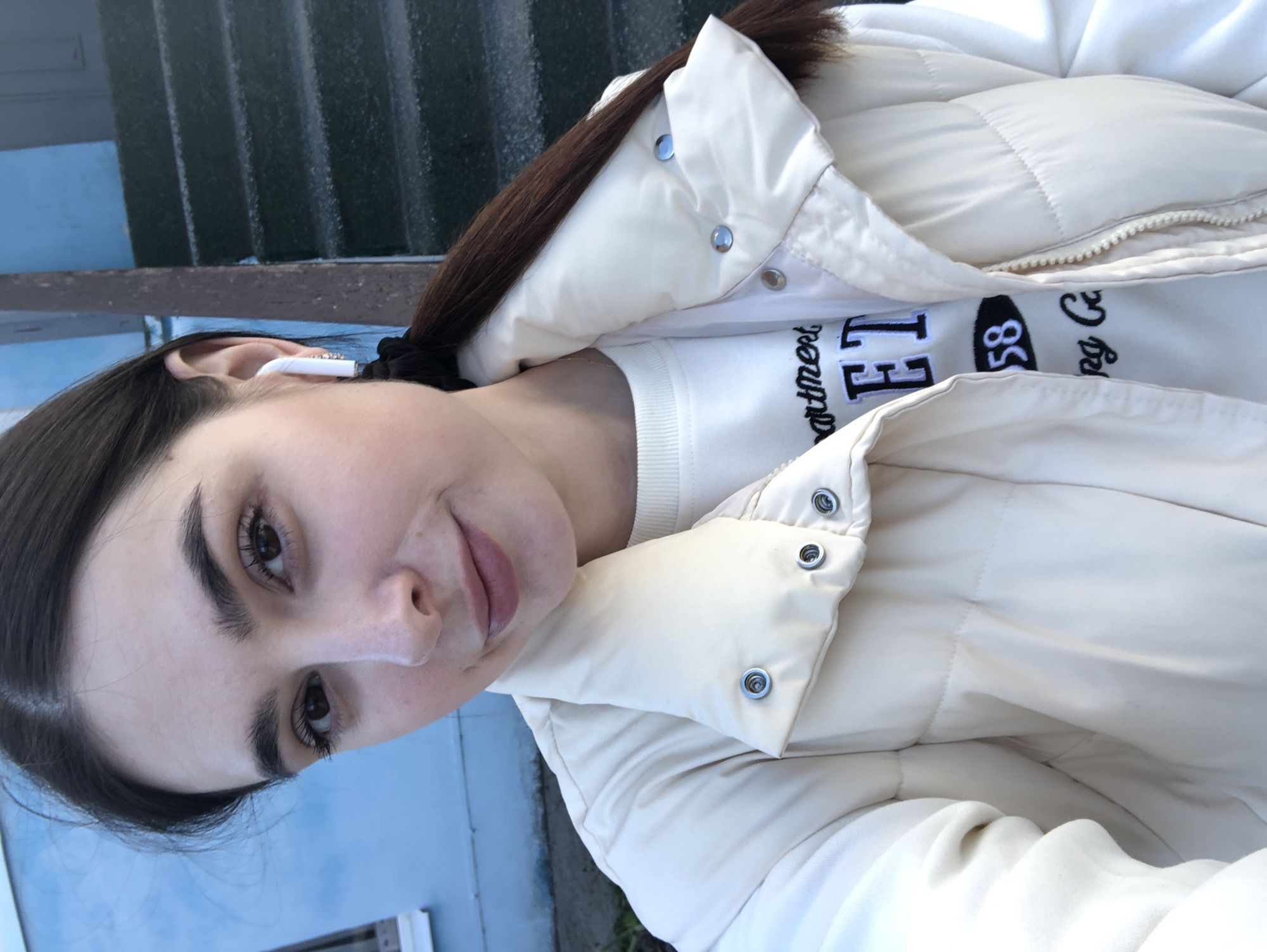 A young woman with dark brown hair pulled back in a ponytail, wearing a white puffer jacket over a white sweater with the words "Department El 58" on it, is taking a selfie. She is wearing a white earbud in her left ear. The background shows a building with dark gray siding and a blue-painted lower level. Based on the information provided, the location is the city of Chaikovsky, Russia.