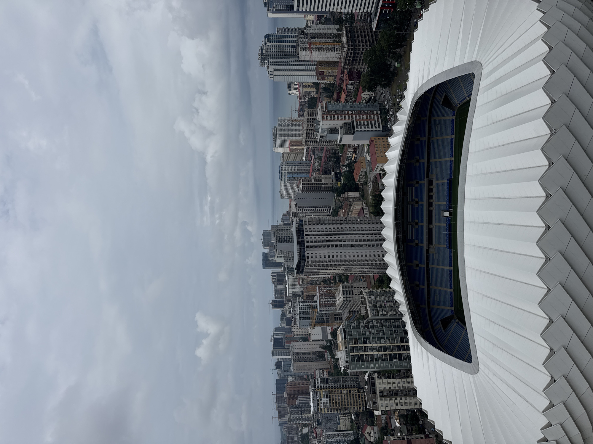 An aerial, wide-angle shot captures the Batumi Stadium in Batumi, Georgia, under a cloudy, overcast sky. The foreground is dominated by the stadium's distinctive white, wave-like roof structure, which extends across the lower portion of the image. Beyond the stadium, a dense urban landscape unfolds, characterized by numerous high-rise apartment buildings and other structures. The buildings vary in height, color, and architectural style, creating a complex cityscape. In the distance, the Black Sea is visible, forming a hazy blue line on the horizon, further emphasizing the coastal location of the city. The overall atmosphere is one of a sprawling, modern city under a muted, diffused light.