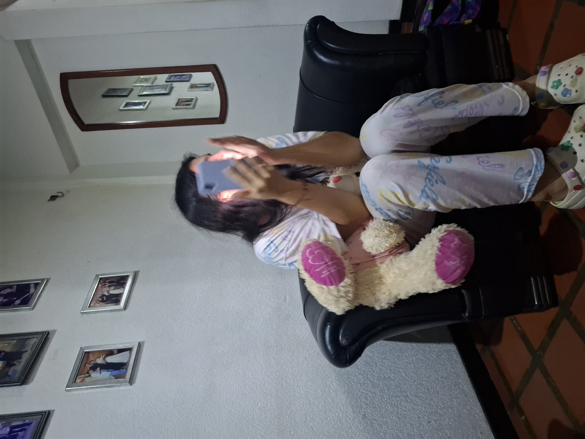 A young person is sitting in a black leather armchair, holding a bright smartphone in their hands. The phone is illuminated, casting light on the person's face, which is obscured by their hair. They are wearing a colorful t-shirt and patterned pajama pants. A plush toy, a beige teddy bear with pink accents, rests on the person's lap. The room features white walls adorned with framed photographs and a tall, oval mirror reflecting several smaller framed pictures. The floor is tiled with reddish-brown squares. This scene is located in Juan Griego, Venezuela.