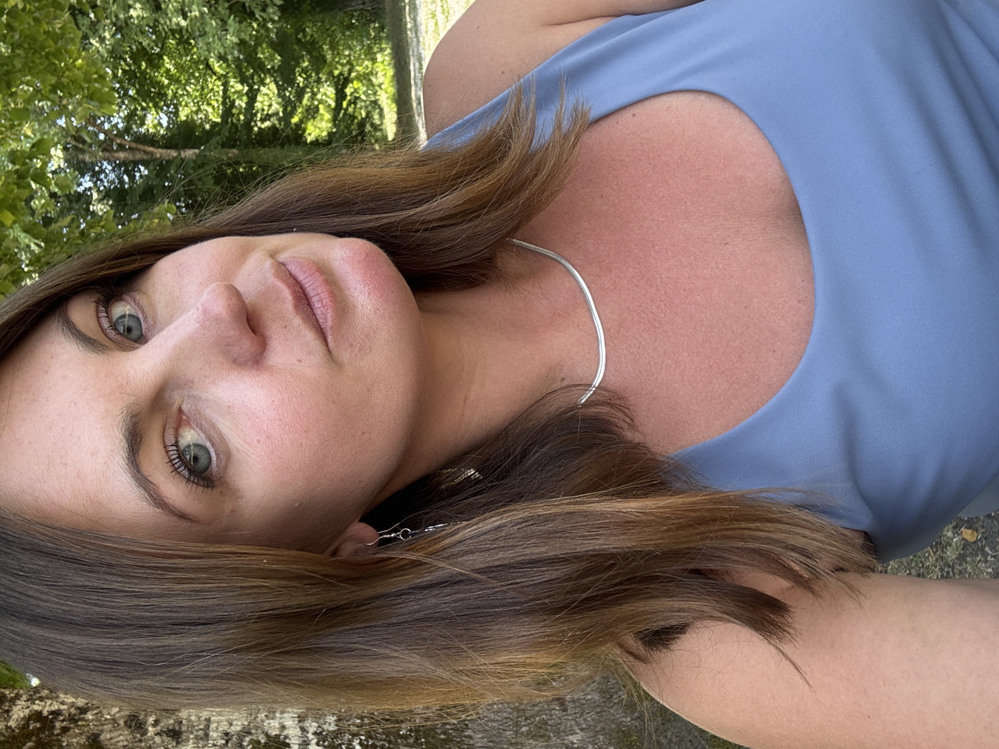 A woman with shoulder-length brown hair and blue-green eyes is looking directly at the camera. She is wearing a blue tank top and a silver necklace. The background is a blur of green trees, suggesting an outdoor setting, possibly in a park or wooded area. The lighting is natural and appears to be daylight. The image does not contain enough information to determine the specific location within Coteaux du Lizon, France.