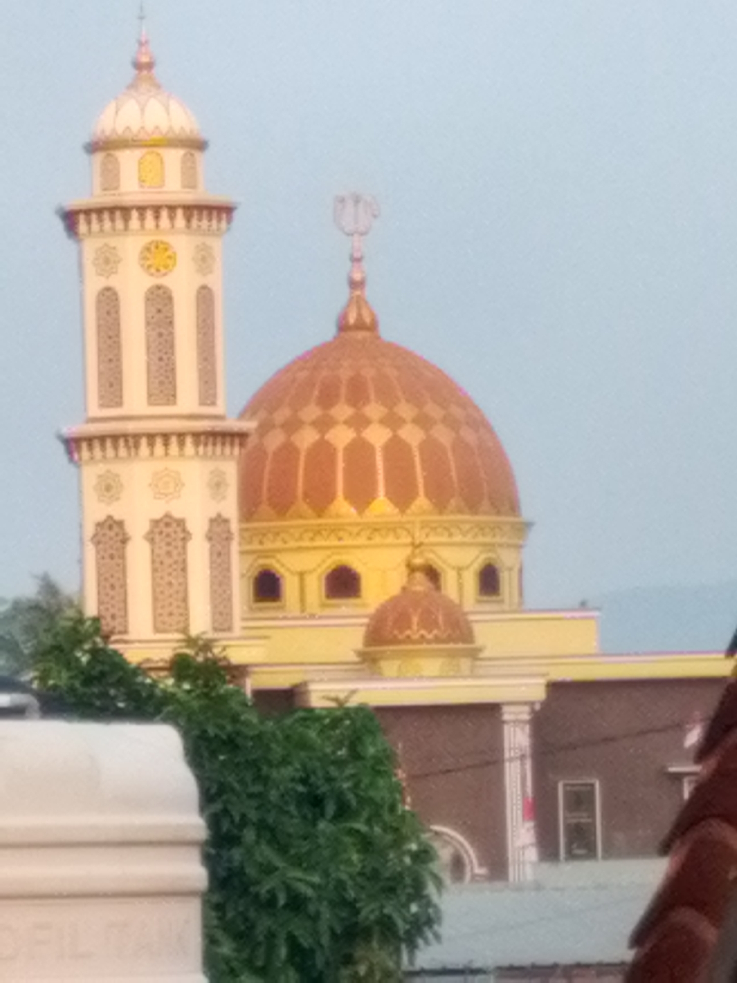 The image shows a mosque with a tall minaret and a large dome. The mosque is painted in yellow and white, with intricate patterns on the minaret. A green tree is in the foreground, partially obscuring a white structure with text. The background is a clear sky. This mosque is identified as Masjid Baiturrahman located in Bandar Lampung, Indonesia, a prominent landmark in the city.