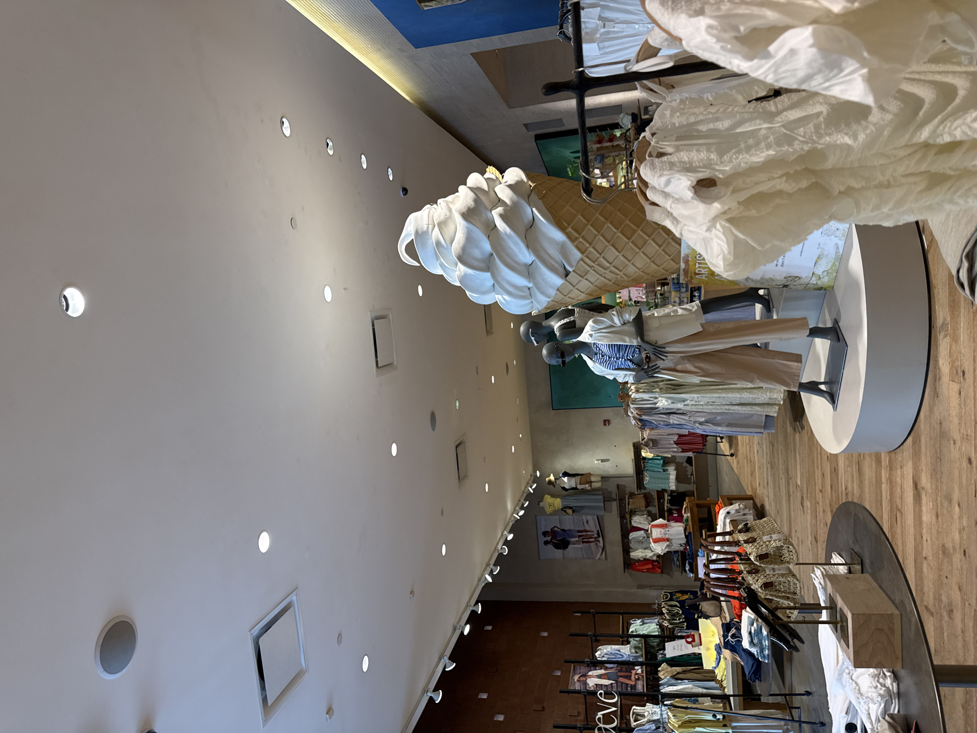The image captures the interior of an Anthropologie store. A large artificial ice cream cone prop is prominent in the foreground. There are mannequins displaying clothing. Racks of clothing are arranged throughout the store. The store is lit by recessed ceiling lights. This location is in Austin, United States of America.