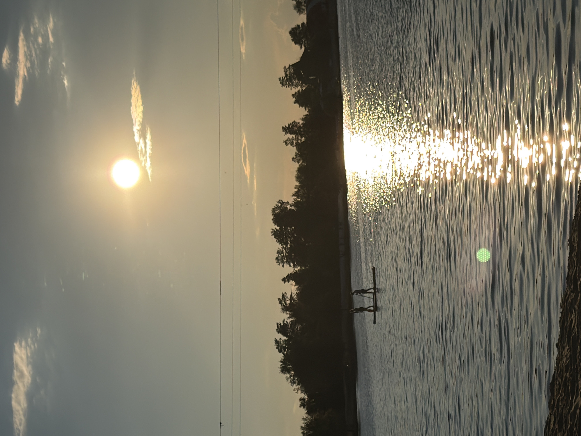 The image depicts a serene outdoor scene at dusk, likely on a lake or calm body of water. Two silhouetted figures are visible standing on a paddleboard, each holding a paddle. They are positioned in the middle of the water, facing away from the camera. The sun is setting, casting a warm, golden glow across the sky and reflecting brilliantly on the water's surface, creating a shimmering path of light. In the background, a line of trees forms a dark silhouette against the twilight sky, with a few buildings or structures faintly visible amongst them. Overhead, a thin power line stretches across the frame. The foreground shows a sliver of sandy shore. The overall atmosphere is peaceful and tranquil, capturing a moment of leisure at the end of the day.