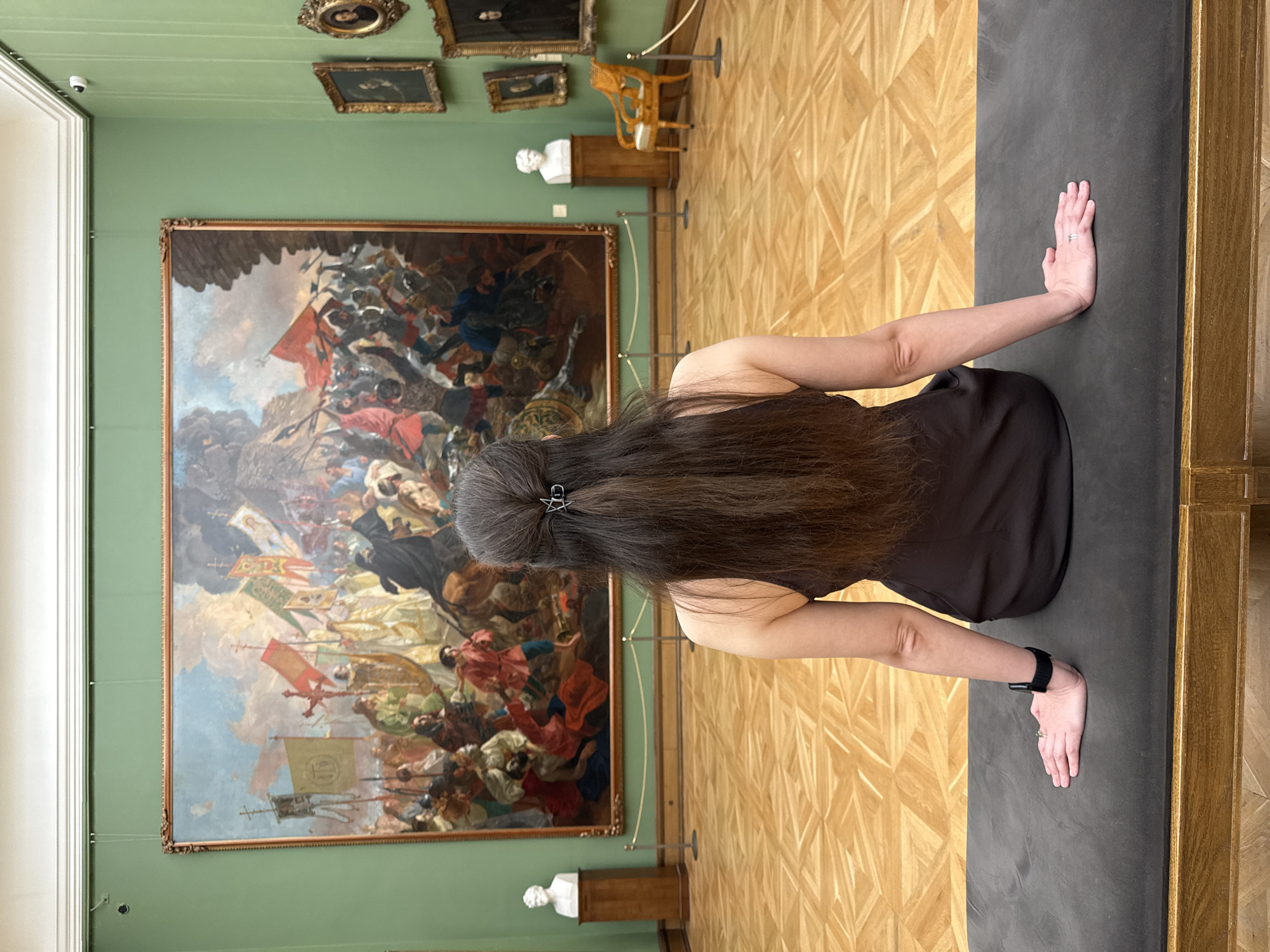 A woman with long brown hair, held back with a star-shaped hair clip, stands in an art gallery. She is facing away from the camera with her hands pressed against a grey surface. The gallery has parquet flooring and light green walls. A large, framed painting depicting a historical battle scene hangs on the wall behind her. Smaller paintings, a bust on a pedestal, and a decorative chair are visible in the background. The location is most likely a museum in Moscow, Russia, given the style of art and architecture.