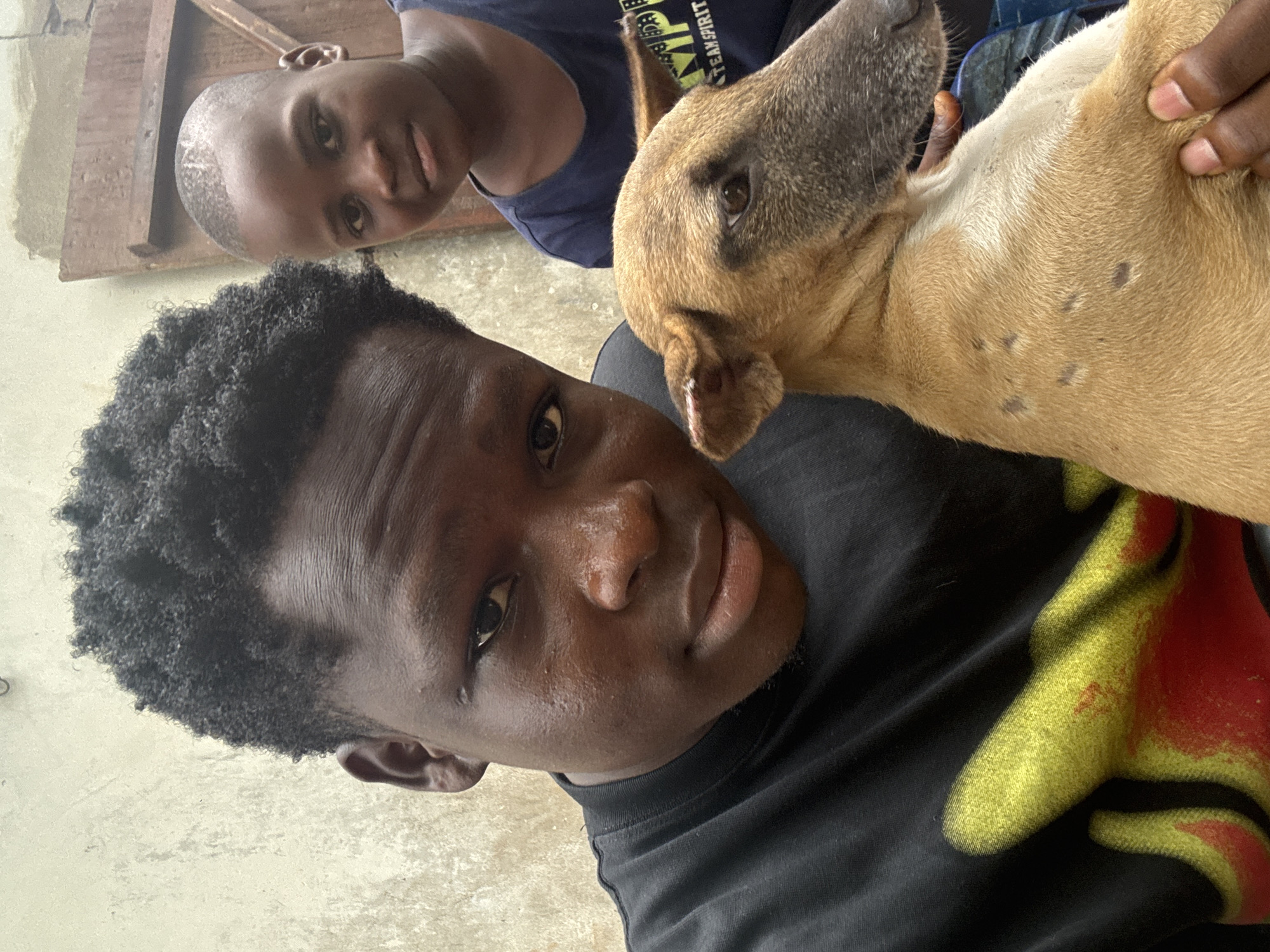 Aboh, Nigeria. A medium shot captures two Black males and a dog in an outdoor setting. The primary subject, in the foreground, is a dark-skinned Black male with short, curly hair. He is wearing a black t-shirt featuring a faded yellow and red design. He is looking directly at the camera. Behind him and slightly above is a younger, light-skinned Black male wearing a dark blue t-shirt with white lettering. The second male looks toward the camera, slightly to the left. A tan and brown dog with dark spots on its neck is in the right foreground, its head in the primary subject's lap. The background appears to be a concrete or stone surface and a wooden structure. The lighting suggests it is daytime.