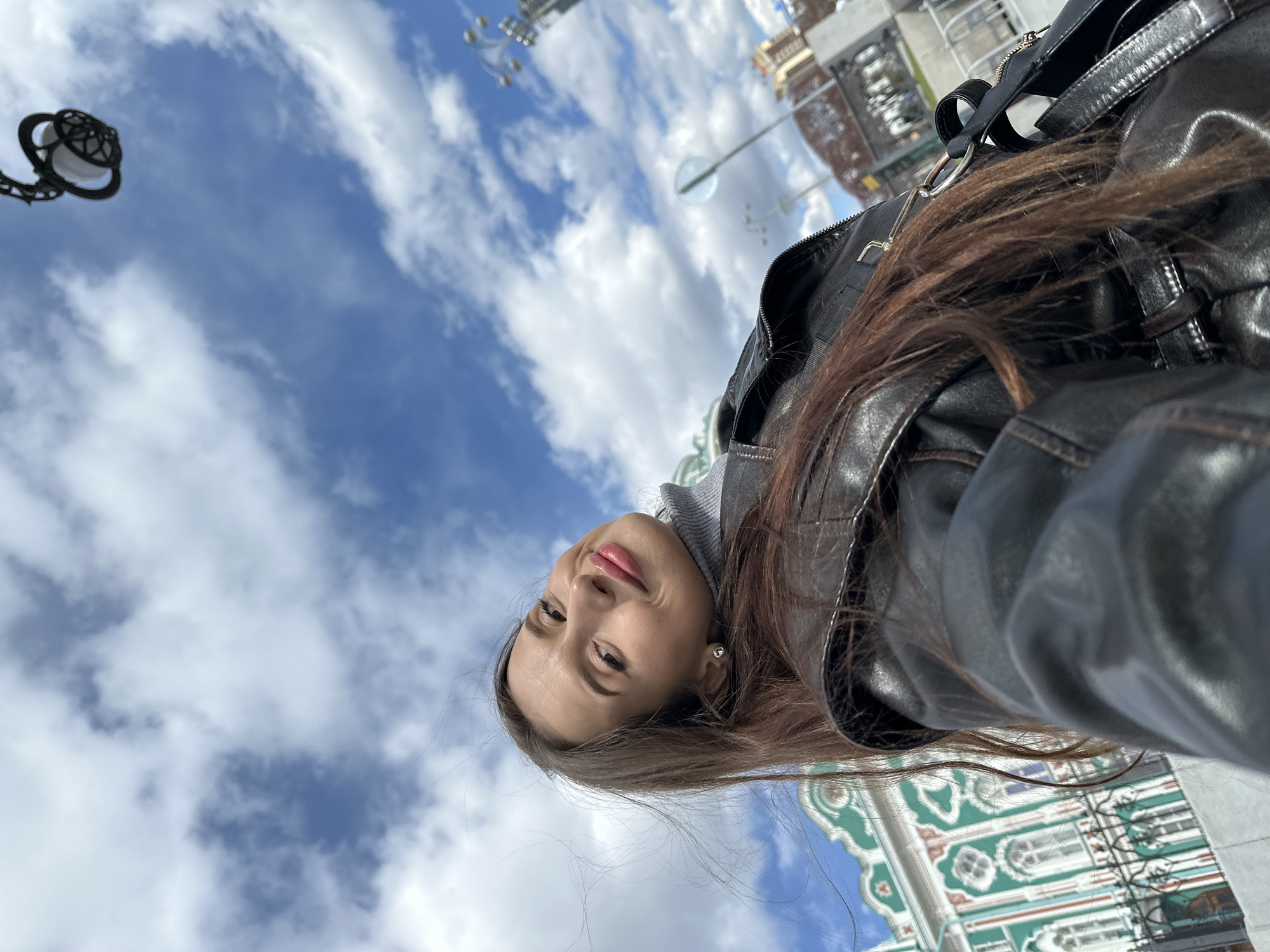 A selfie of a woman wearing a leather jacket and a gray sweater. She is facing the camera with a slight smile. In the background is a blue sky with scattered white clouds. Visible on the right and lower sections of the image are architectural details of the facade of the Cathedral on the Blood in Yekaterinburg, Russia. A lamppost is visible in the upper left corner. Other street lamps can be seen in the background. The photo appears to be taken on a sunny day.