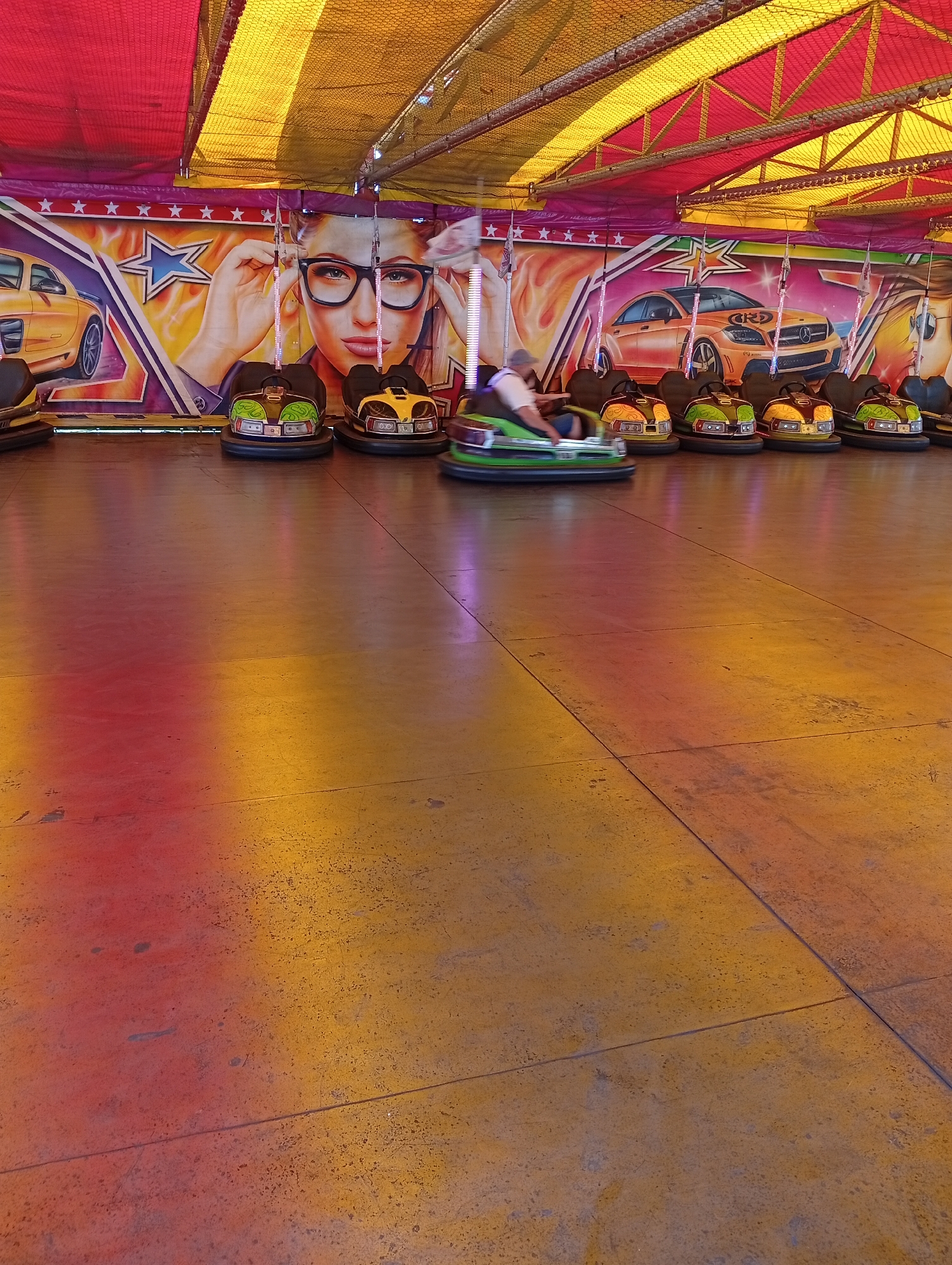 This image depicts an indoor bumper car attraction located in Popovo, Bulgaria. The overhead structure consists of a tent canopy with alternating red and yellow fabric panels supported by a metal truss framework, beneath which a dark mesh netting is visible. The back wall features an extended mural. The mural prominently displays two luxury sports cars, one yellow and one orange-brown, both resembling Mercedes-Benz C63 AMG models, accompanied by stylized star emblems. Between the cars, a large portrait of a woman with dark hair and black-rimmed glasses is shown, with her hands adjusting the frames.
The floor of the arena is a flat, reflective surface, likely metal, exhibiting color gradients of red, orange, and yellow, which appear to be reflections from the overhead lighting and canopy. Several dark lines on the floor indicate panel seams or tracks. Approximately ten bumper cars are present. Most cars are stationary, positioned along the mural wall, with various designs in yellow, green, and black. One green bumper car in the lower-left to mid-left portion of the frame is in motion, evidenced by slight blur. A person wearing a light-colored shirt and a cap is operating this moving bumper car. Each bumper car is equipped with a vertical pole extending upwards, connecting to the power grid (not visible).