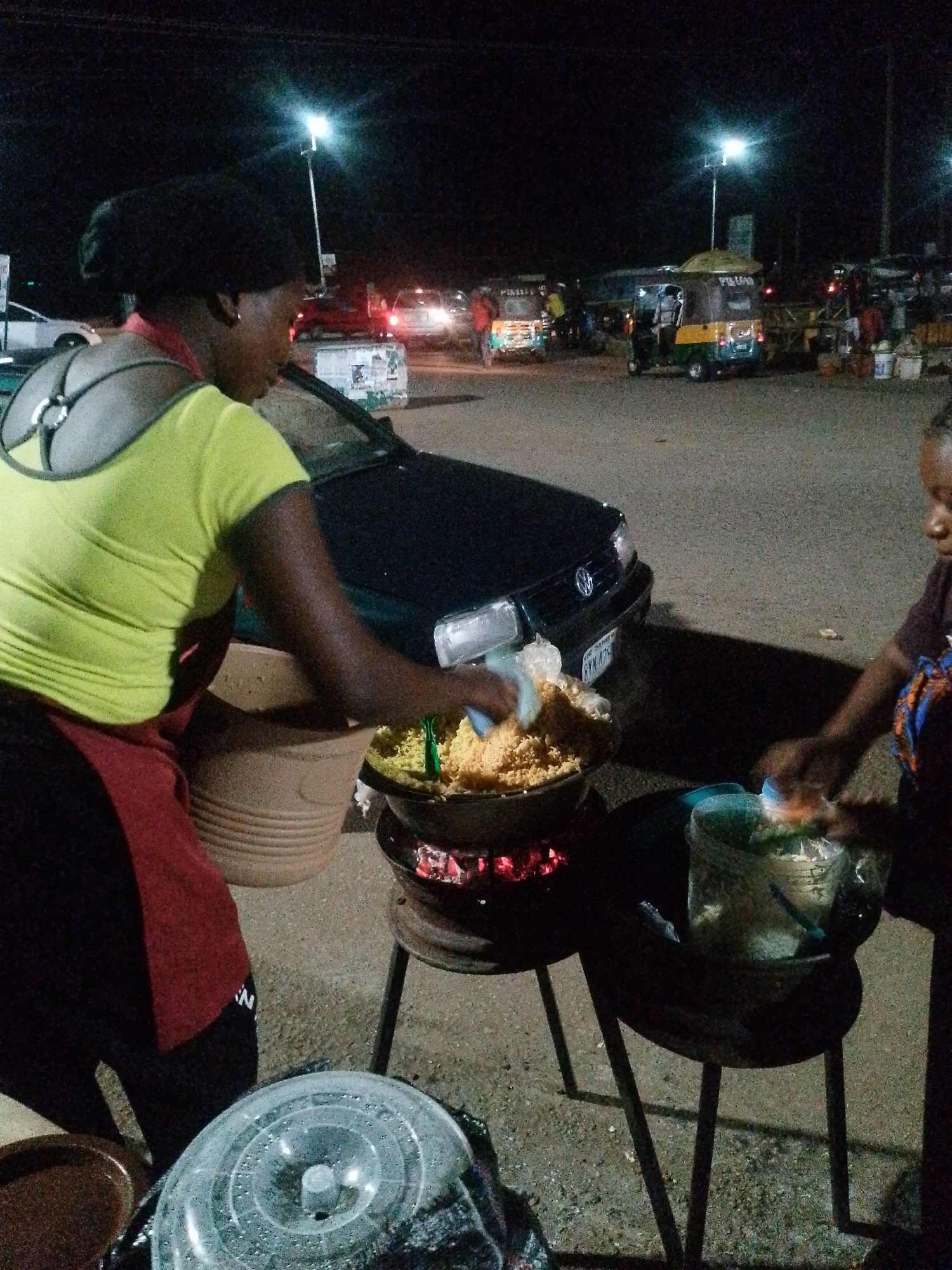In the city of Bukuru, Nigeria, at nighttime, two women are engaged in food preparation near a roadside stall. The woman on the left, wearing a bright yellow shirt and a red apron, is stirring a large pot of food placed over glowing embers. Beside her, a large clay pot rests on the ground. To the right, another woman, partially visible, is also tending to food preparation, with a plastic container of ingredients in front of her. Several vehicles, including a dark-colored car and a colorful auto-rickshaw, are parked in the background, suggesting a busy street or market area. The scene is illuminated by artificial lights.