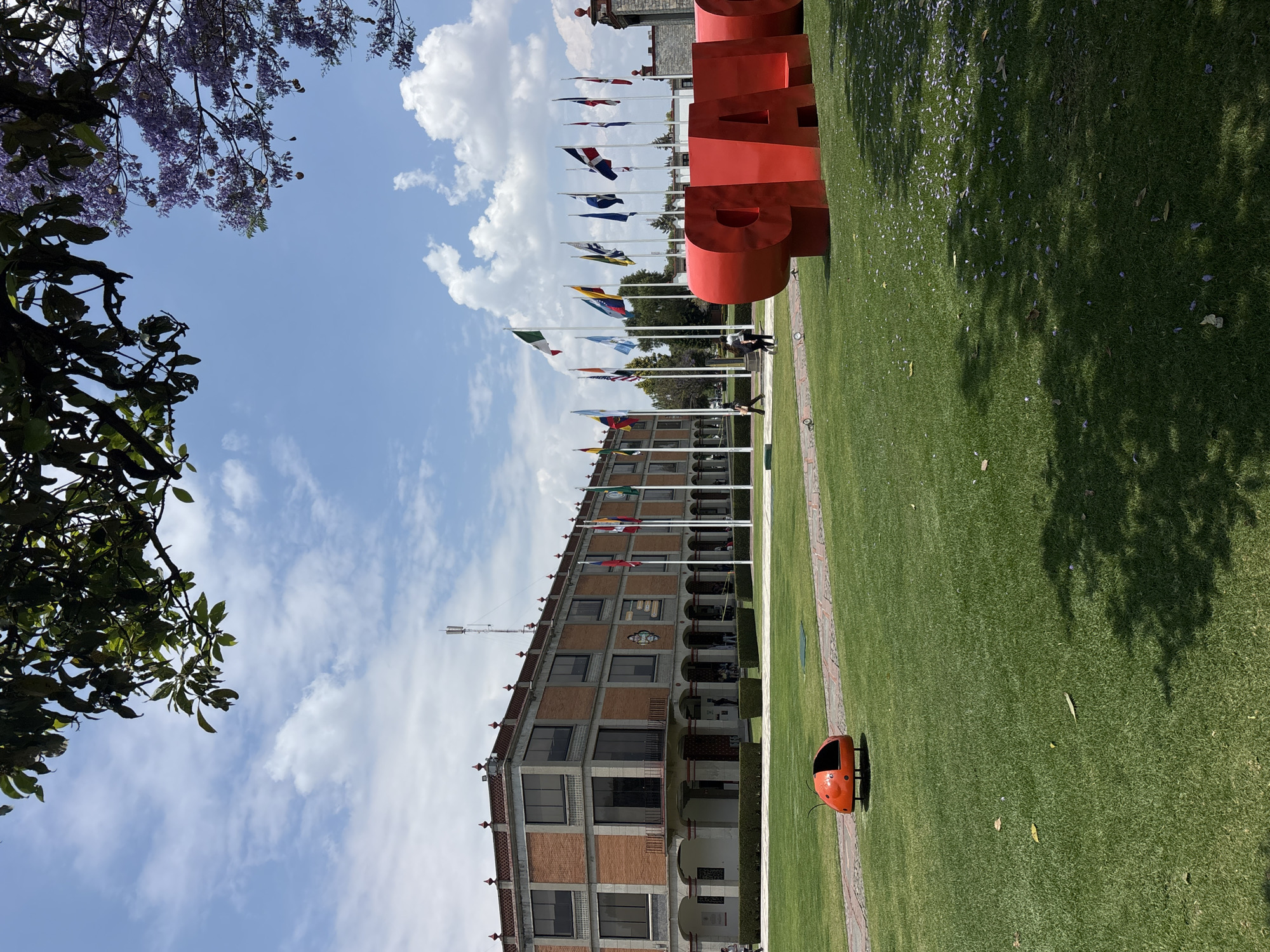 The image depicts a grassy area at the Universidad de las Américas Puebla (UDLAP) in San Andrés Cholula, Mexico. A row of flags on poles stands in front of a building constructed of brick and concrete with arched windows. In the foreground, large red letters spell out "UDLAP." A red ladybug-shaped trash can sits on the grass. The sky is partly cloudy, and tree branches with foliage are visible on the left side of the image. Scattered purple flower petals are visible on the grass. Two people stand between the flags.