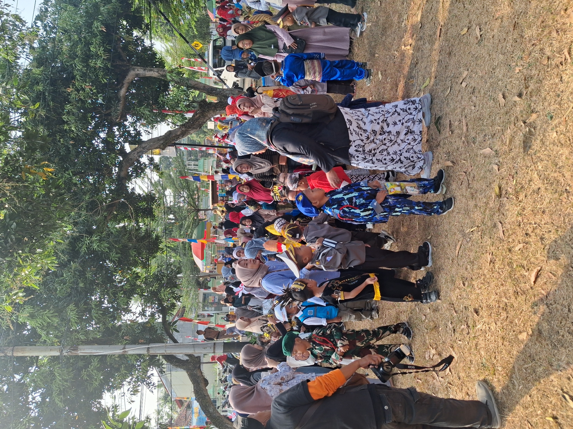 This is a full-shot, eye-level, outdoor photograph taken in Balapulang, Indonesia, likely during a children's parade or event, given the costumes and festive decorations. The scene is set on a dry, grassy ground scattered with fallen leaves, under the dappled shade of large trees with lush green foliage. In the background, several Indonesian flags and colorful banners are visible, hinting at a national celebration.
The foreground is dominated by a crowd of people, both adults and children. Many children are dressed in various costumes, including what appear to be military uniforms (camouflage, blue uniforms with badges), traditional attire, and a boy in a blue and gold outfit resembling a prince or royal guard. An adult in the left foreground wears dark pants and an orange shirt, holding a camera with a strap. Other adults in the crowd are seen wearing traditional Islamic head coverings (hijabs) in various colors, including beige, brown, and light blue. One woman in the center-right is wearing a light blue striped shirt and a patterned skirt.
The overall atmosphere appears to be one of lively community gathering, with people standing, watching, and interacting. The lighting suggests it is daytime, with sunlight casting distinct shadows on the ground. The presence of a white car in the mid-ground indicates a public space accessible by vehicles.