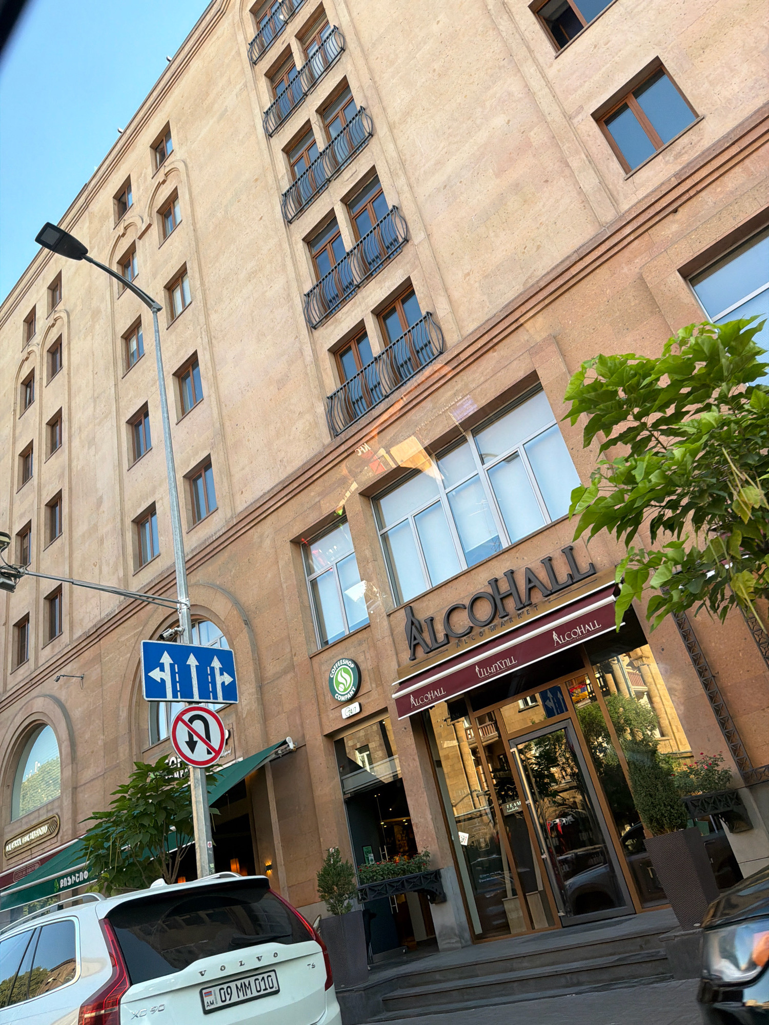 In this eye-level, medium shot, a light beige building with multiple stories, each featuring several windows and decorative balconies, dominates the frame. A street lamp extends from the left side of the image. Below the building, street-level entrances are visible, with the prominent signage "ALCOHALL" appearing above a glass-fronted shop. Several trees and potted plants are present, adding greenery to the scene. Traffic signs, including one with multiple arrows and another indicating no U-turn, are mounted on a pole. A white Volvo XC90 is parked in front of the building. The environment suggests an urban setting, likely a commercial district in Yerevan, Armenia, given the presence of Armenian license plates and the architecture.
