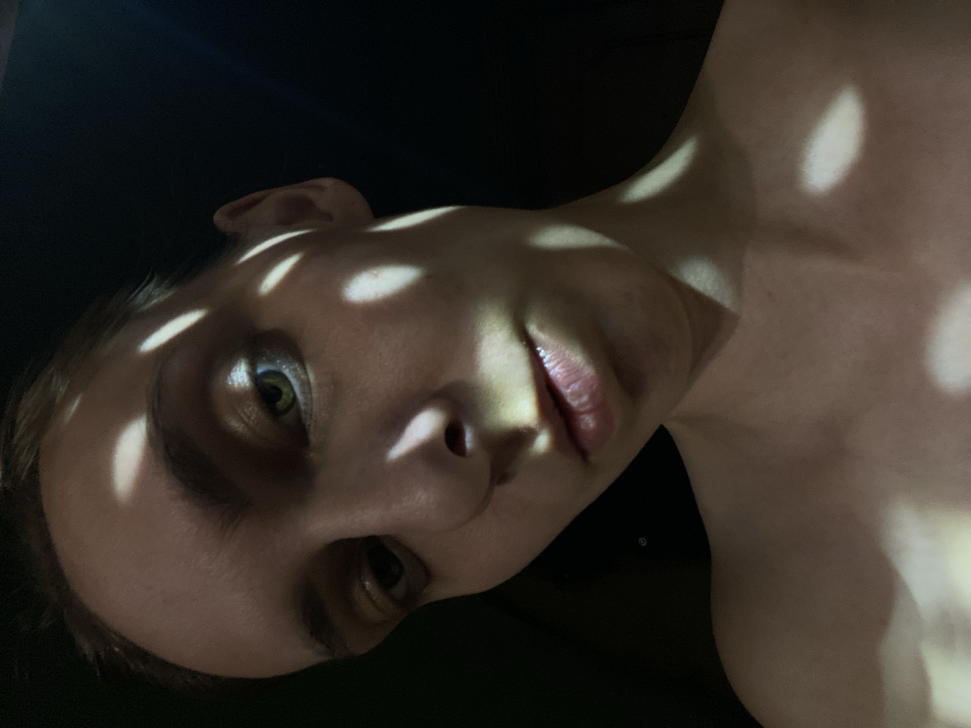 A close-up, low-angle shot depicts a person with light skin in a dimly lit environment. The individual's face and upper body are partially illuminated by dappled light patterns. The person's face is angled towards the camera, with their eyes open and focused. The background is dark and indistinct, obscuring any specific details about the location. There is no verifiable information to suggest the photo was taken in Chaikovsky, Russia.